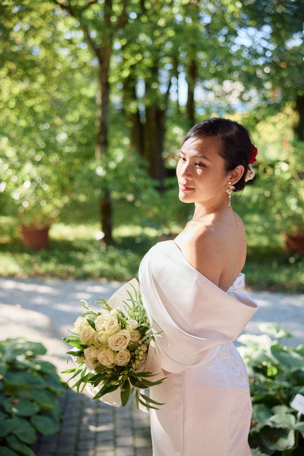 Bride in elegant white dress with cowl neckline holds white floral bouquet in sunlit garden setting.