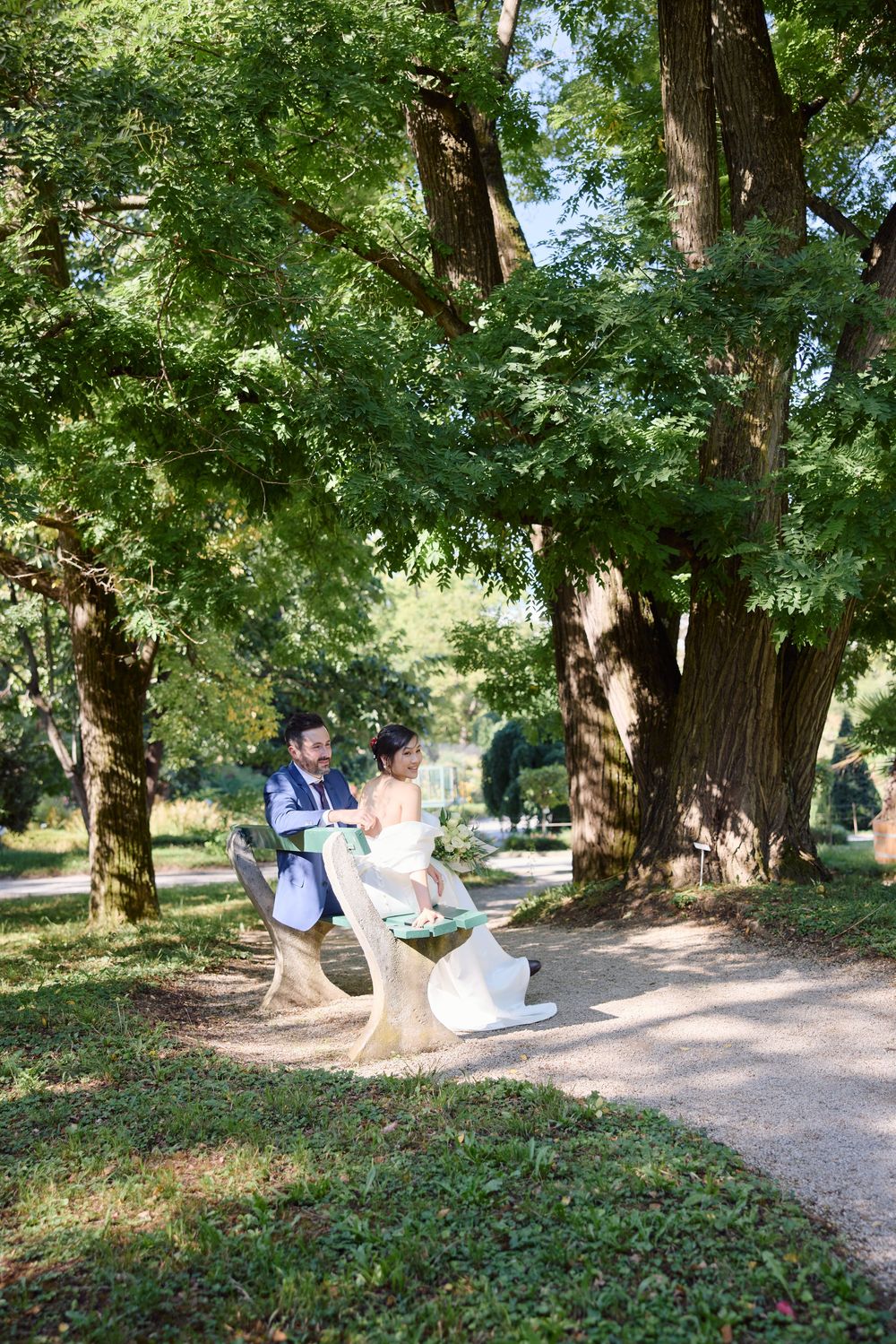 Two people relax on a bench along a peaceful tree-lined gravel path in a tranquil park setting.