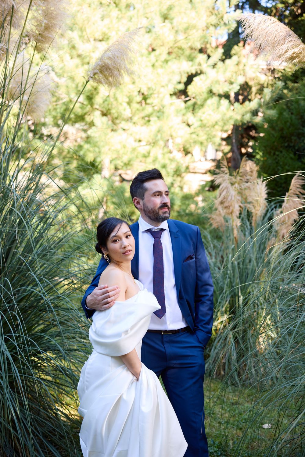 Romantic wedding couple poses together against tall ornamental grasses in a sunlit garden setting.