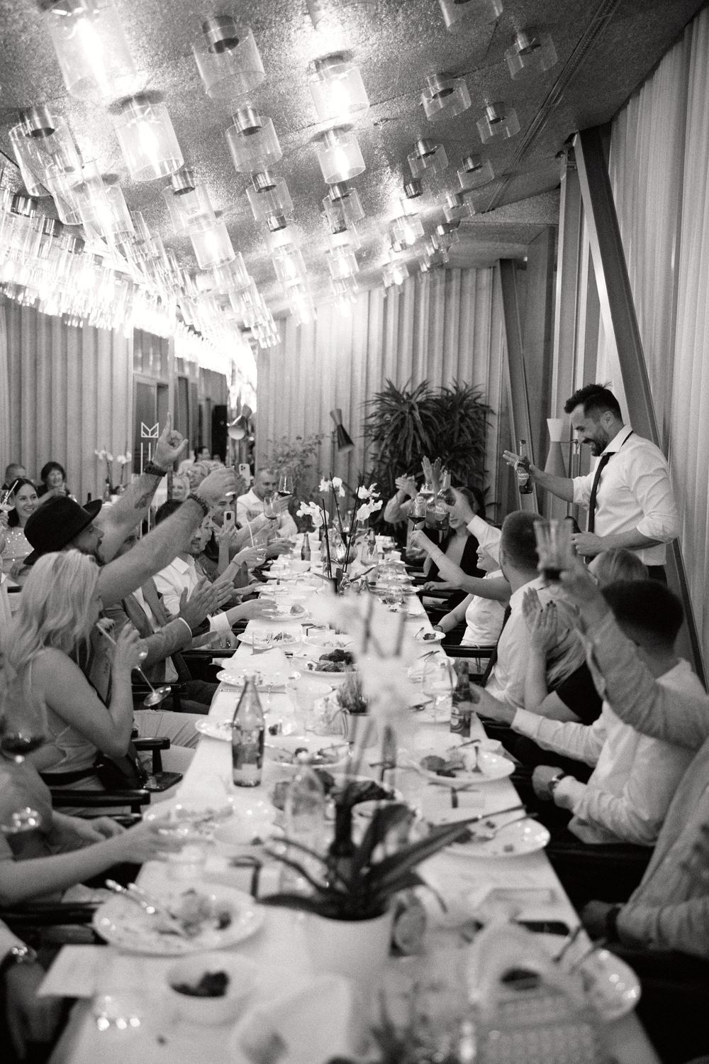 Black and white photo of wedding reception guests raising glasses in celebration under crystal chandeliers.