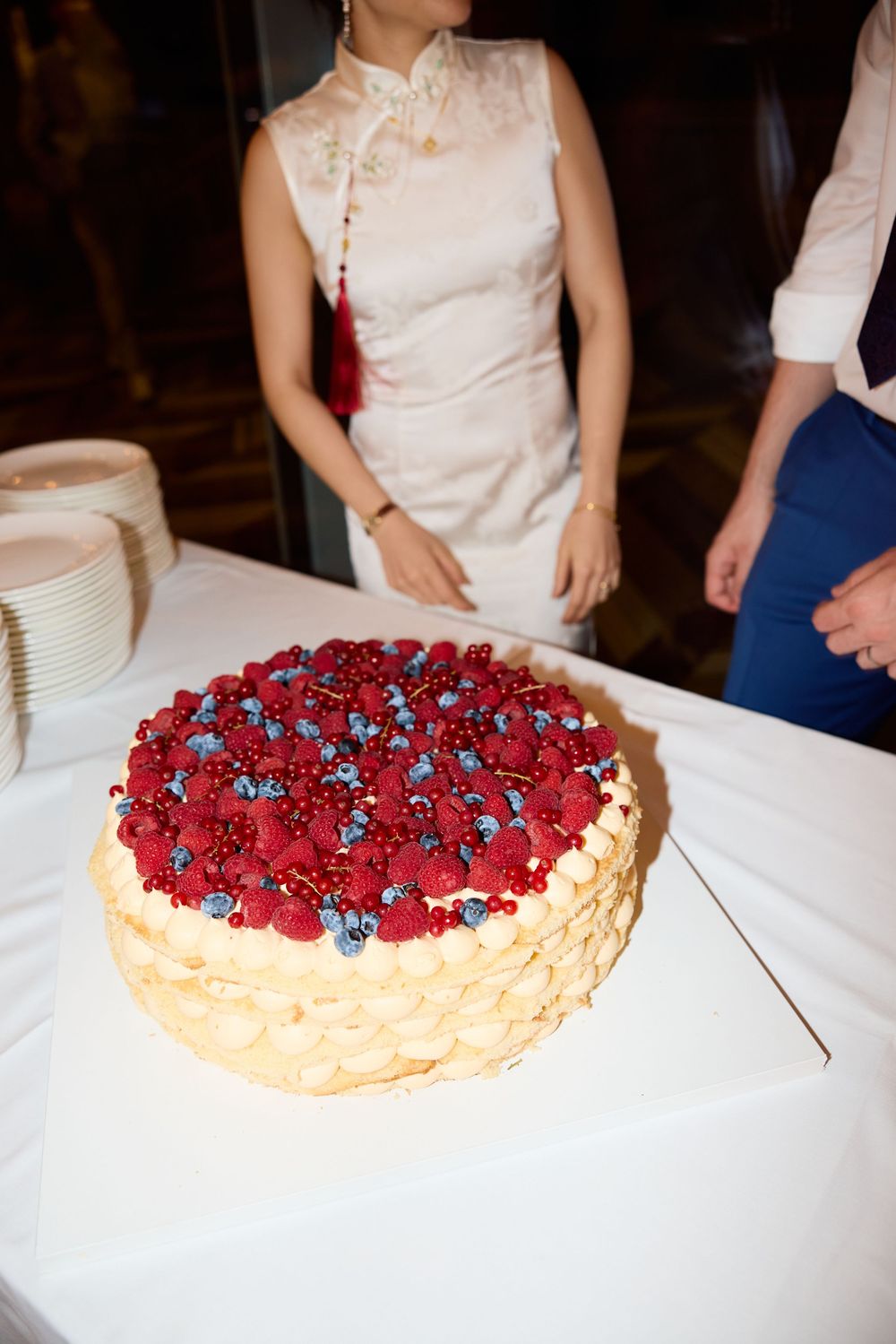 A wedding cake topped with fresh red berries and blueberries is displayed on a white tablecloth.