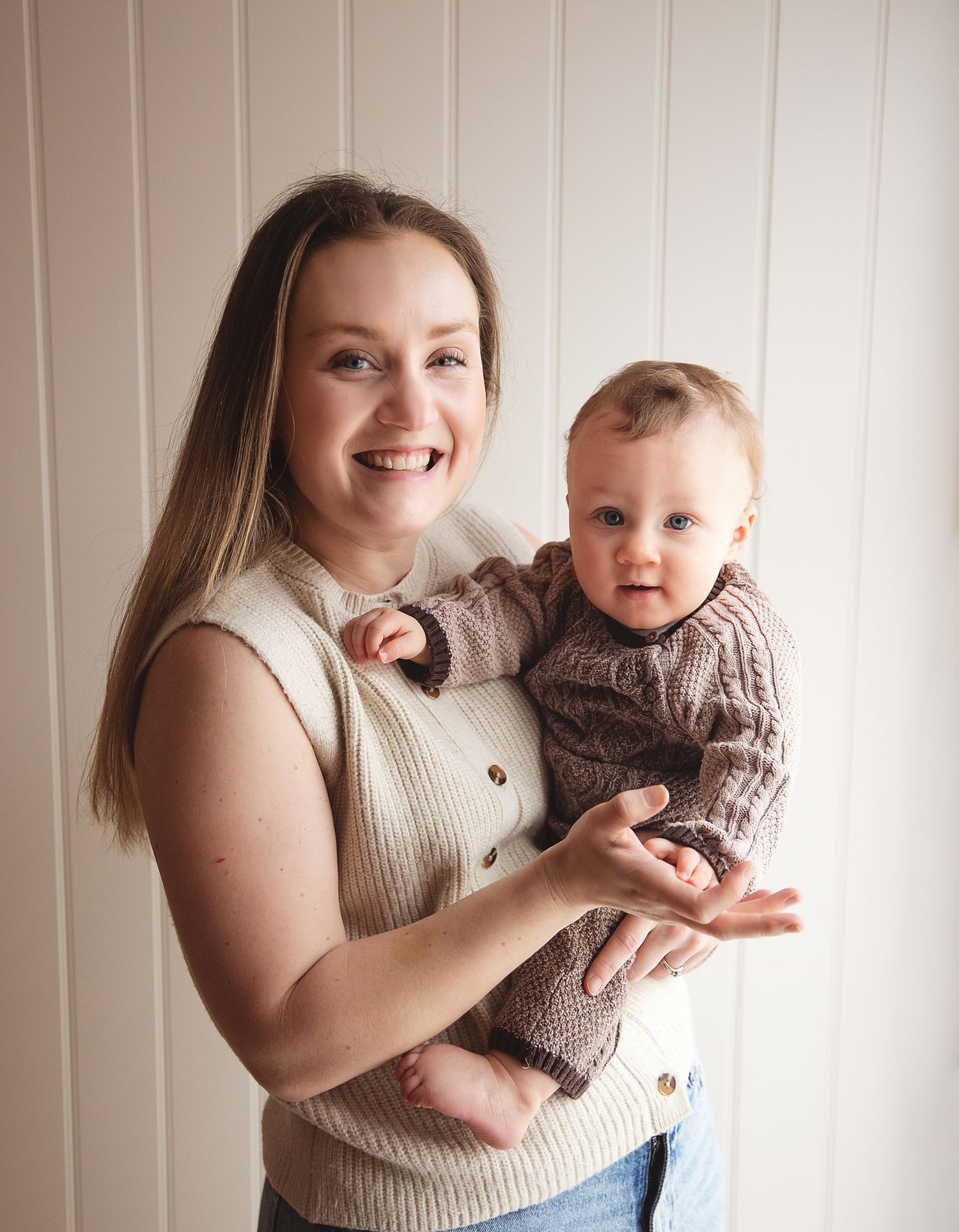 A parent and baby wearing matching brown knit sweaters pose together against a white paneled wall.During a mummy and me photoshoot in Bexley