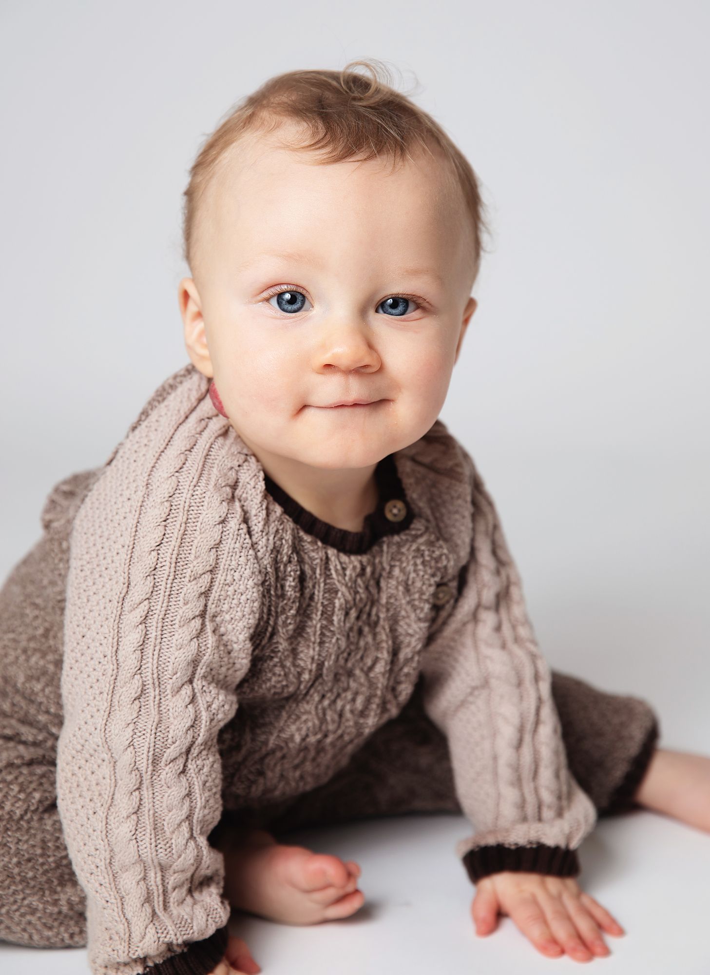Baby wearing a cozy beige cable knit sweater crawls on white background. During a baby photoshoot in Bexley