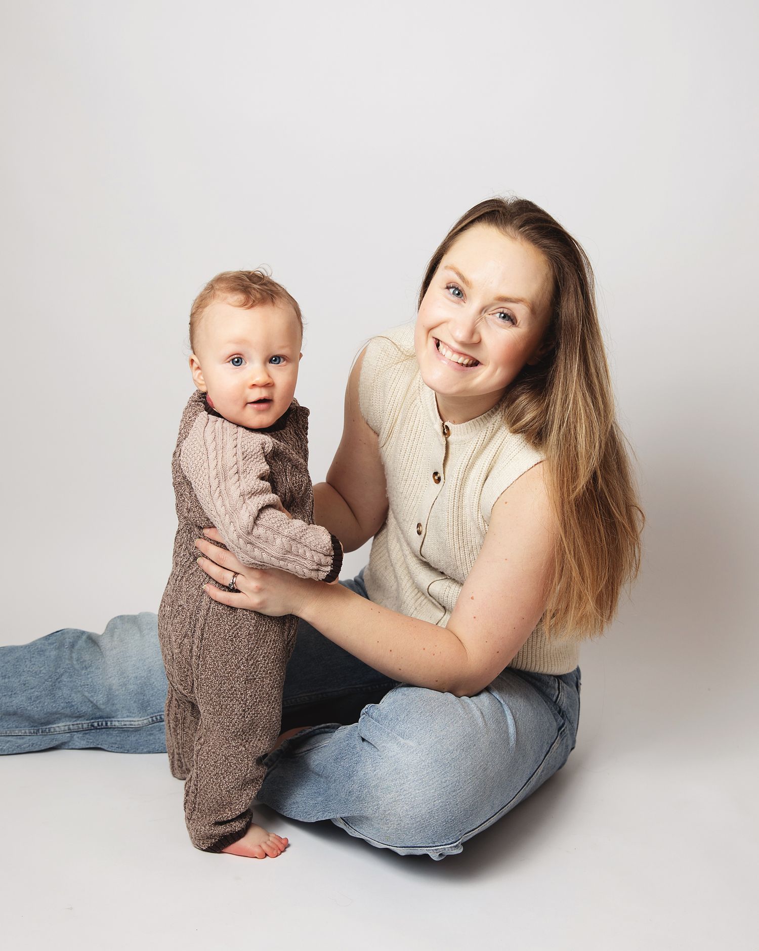 A mother and baby in matching cream sweaters pose together against a white studio backdrop for a series of lifestyle photos in Sidcup