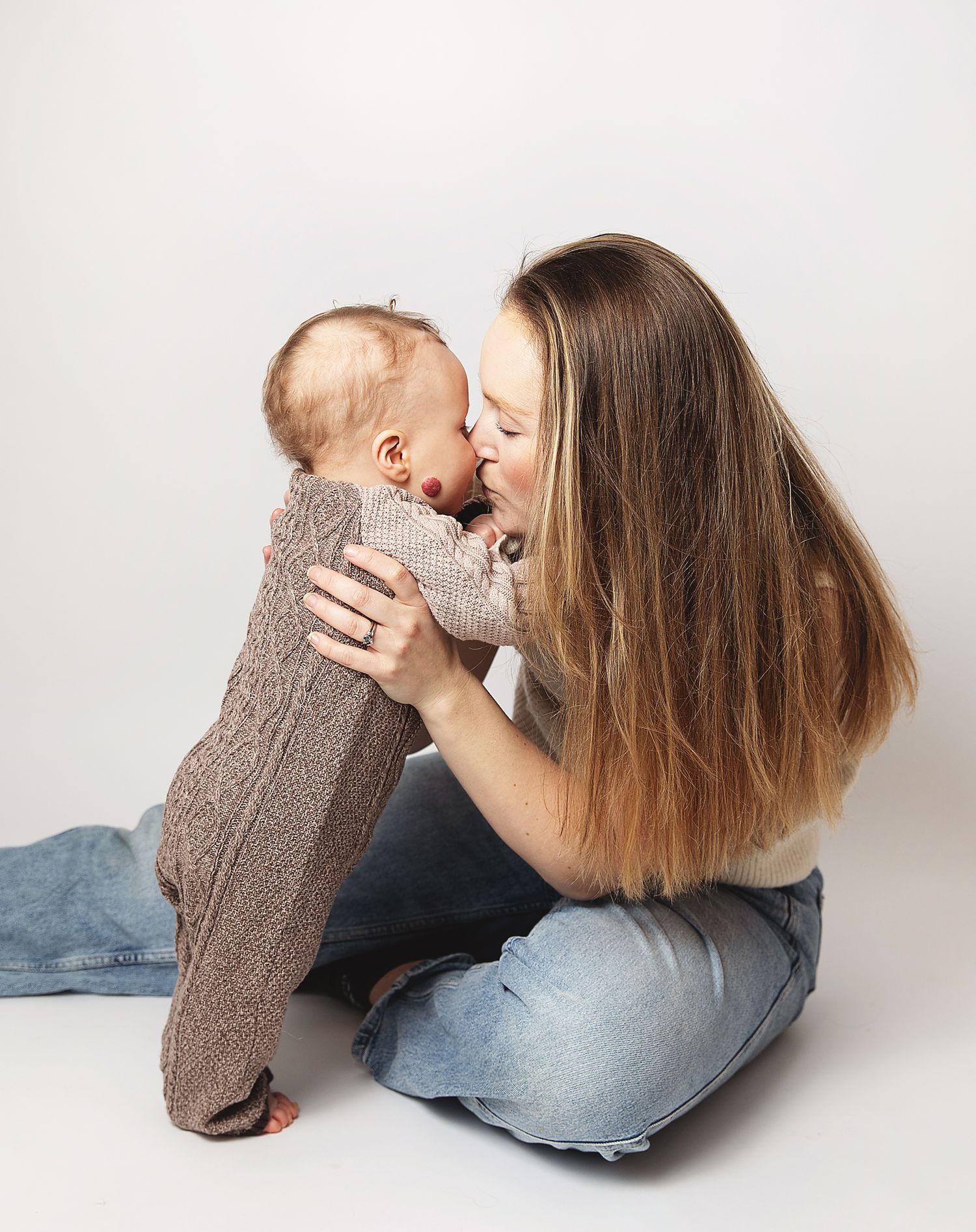 A parent and baby share a tender moment together on a white backdrop while wearing casual clothing. During a baby photoshoot in Bexley