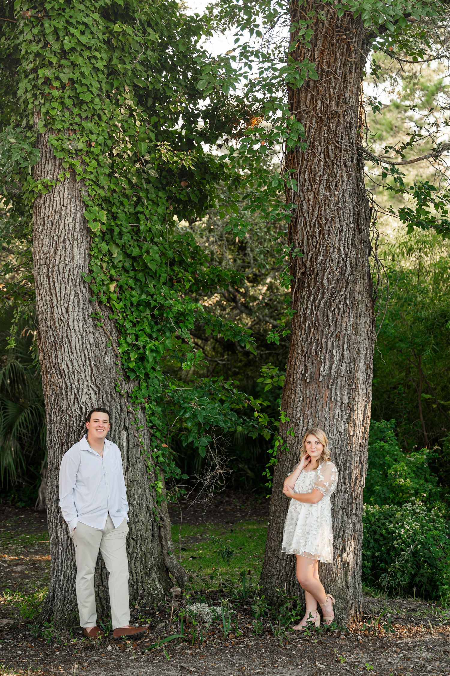A couple poses separately against large tree trunks in a natural outdoor setting.