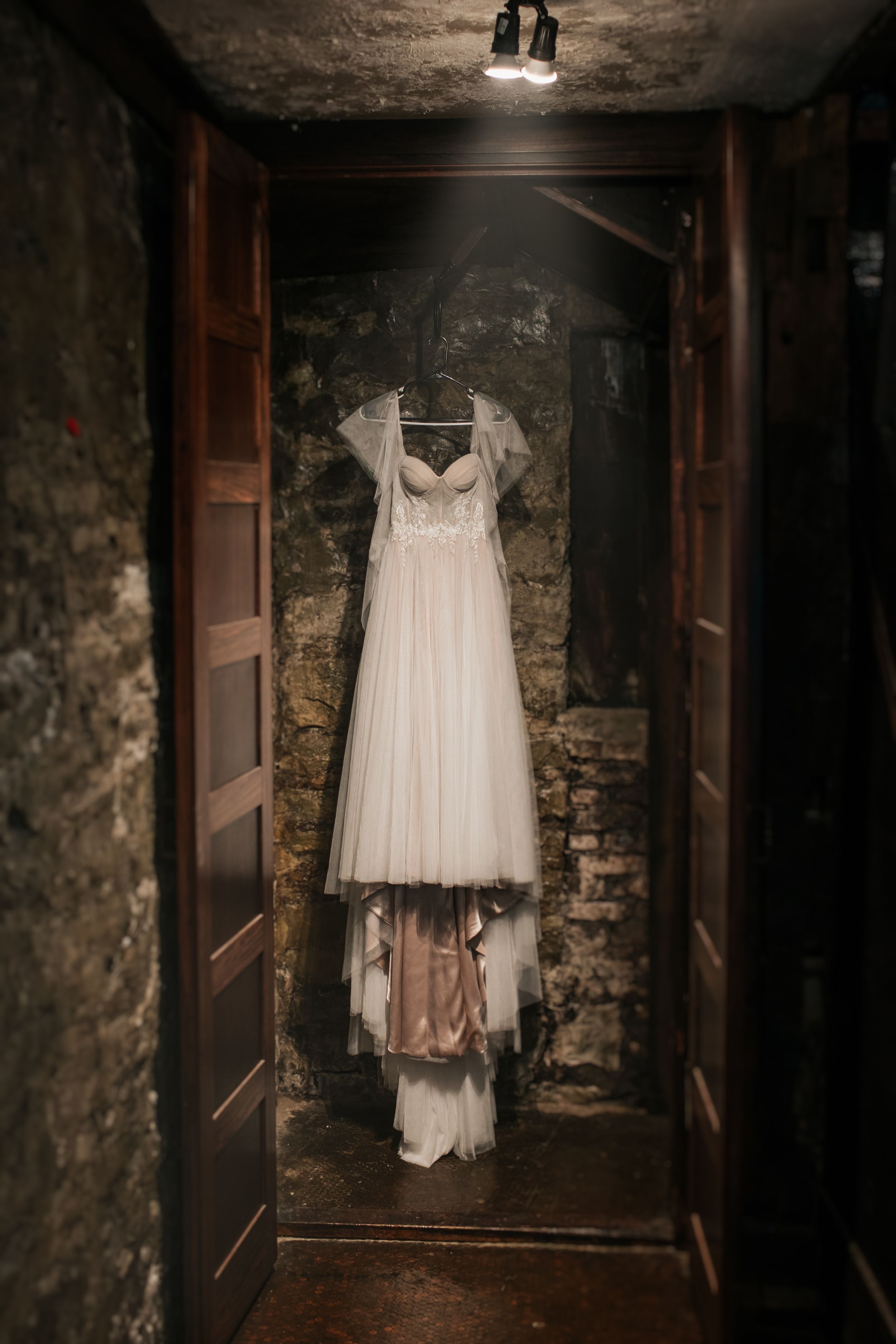 A white wedding dress hangs mysteriously in a dark stone corridor with wooden doors creating an eerie atmosphere.