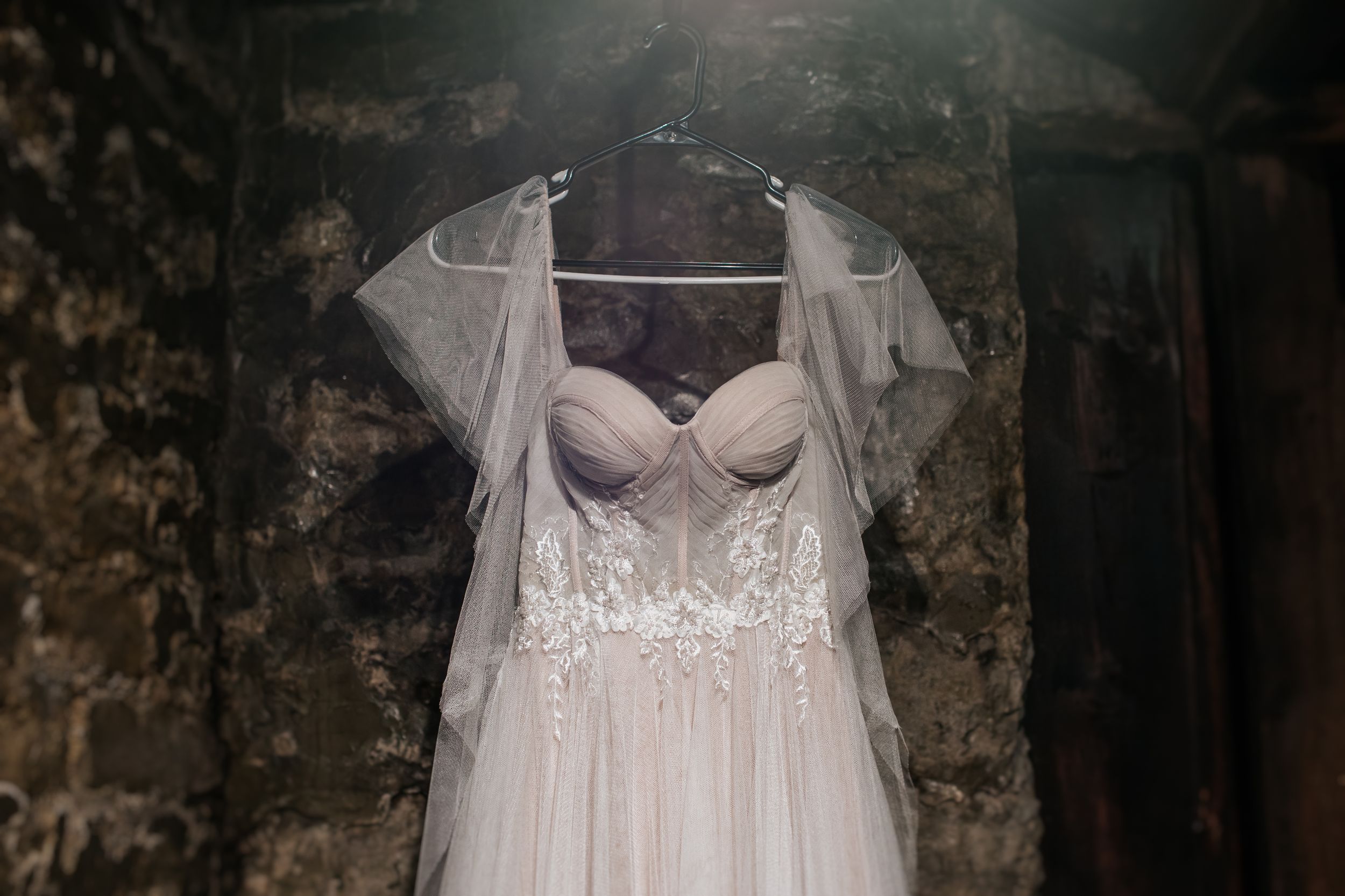 A delicate white wedding dress with lace details and sheer cap sleeves hangs against a rustic stone wall.