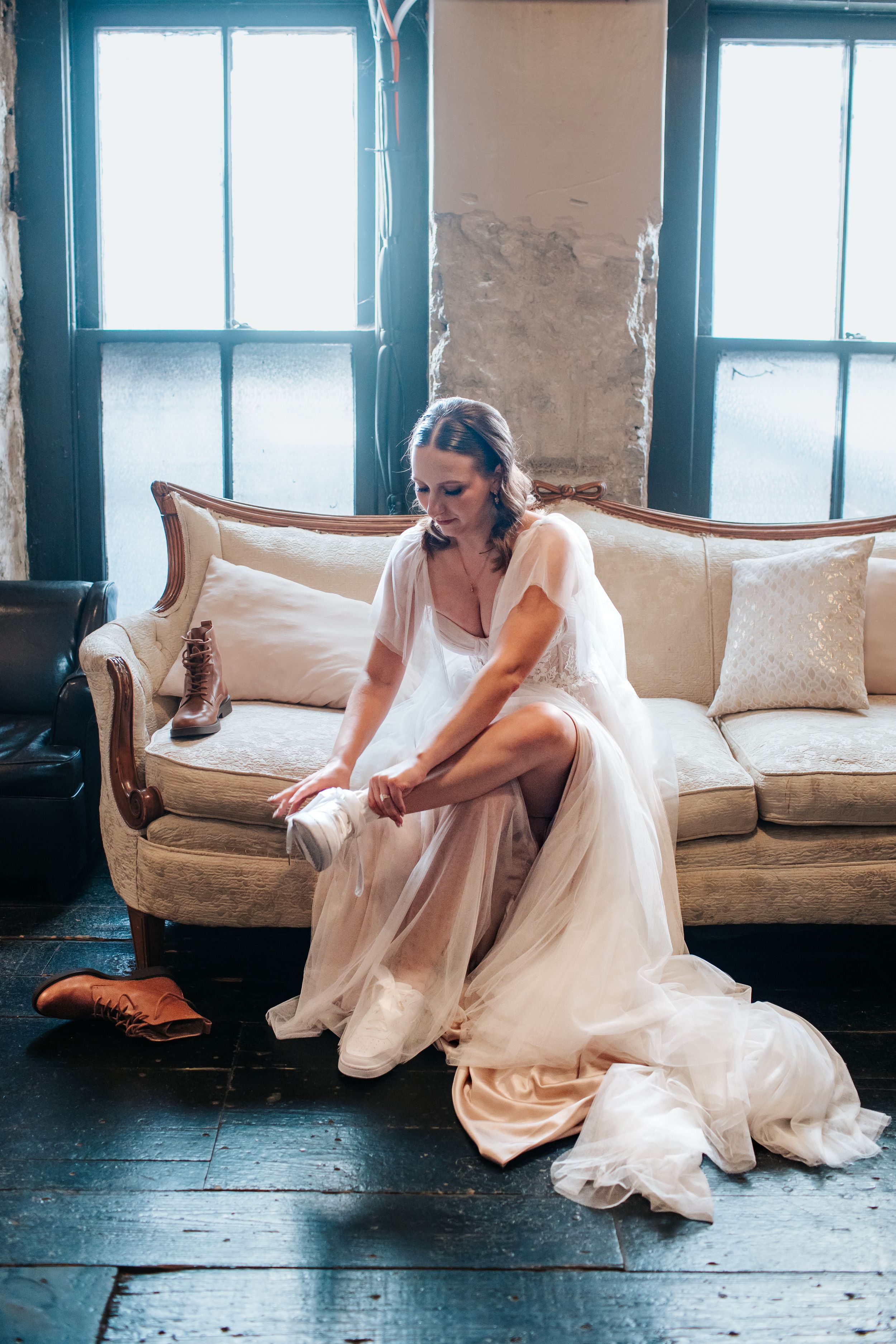 A dreamy bridal scene with flowing white dress and veil on an antique sofa against rustic windows and wooden floors.