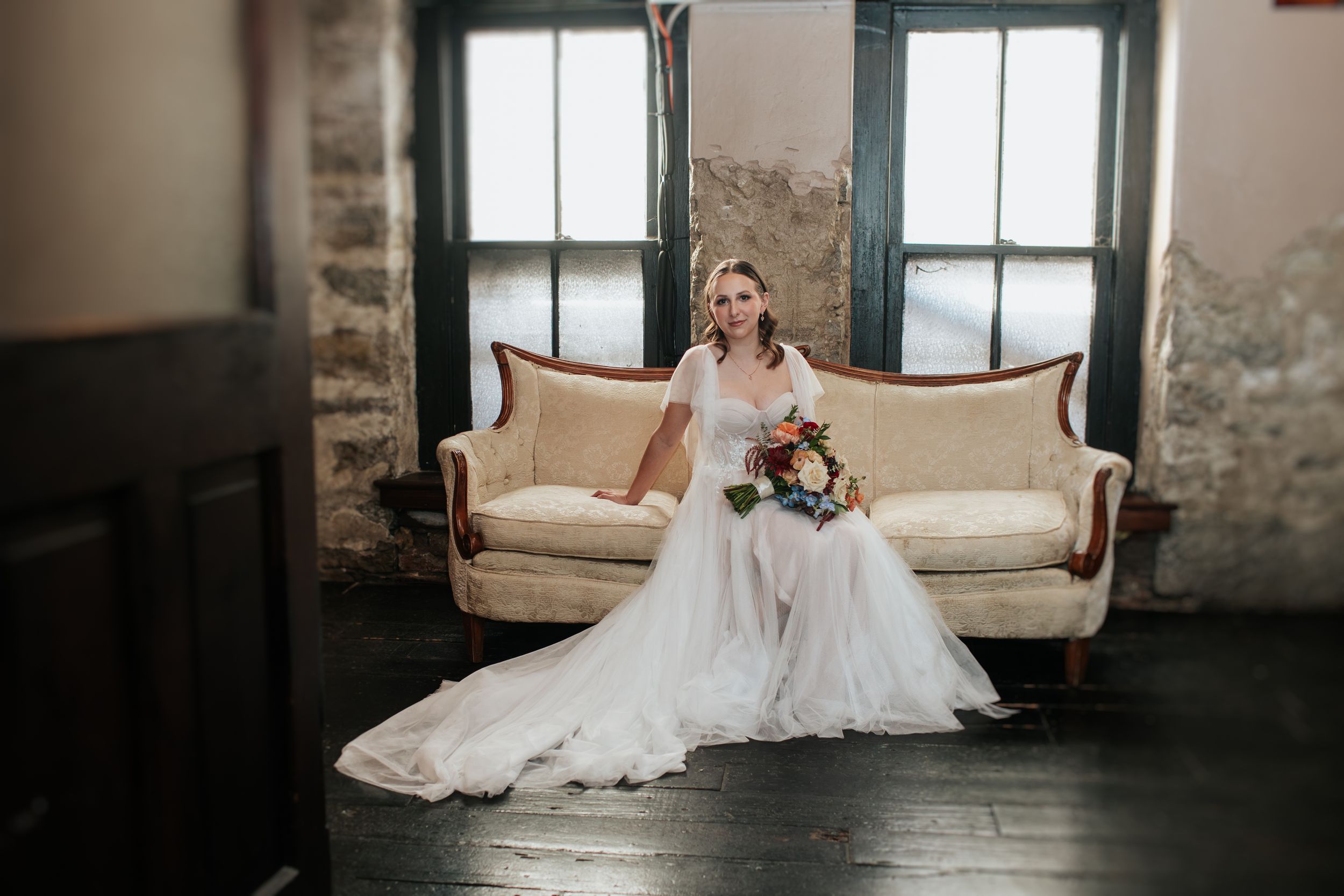 A bride in a flowing white gown reclines gracefully on a cream-colored couch in a rustic stone-walled room.