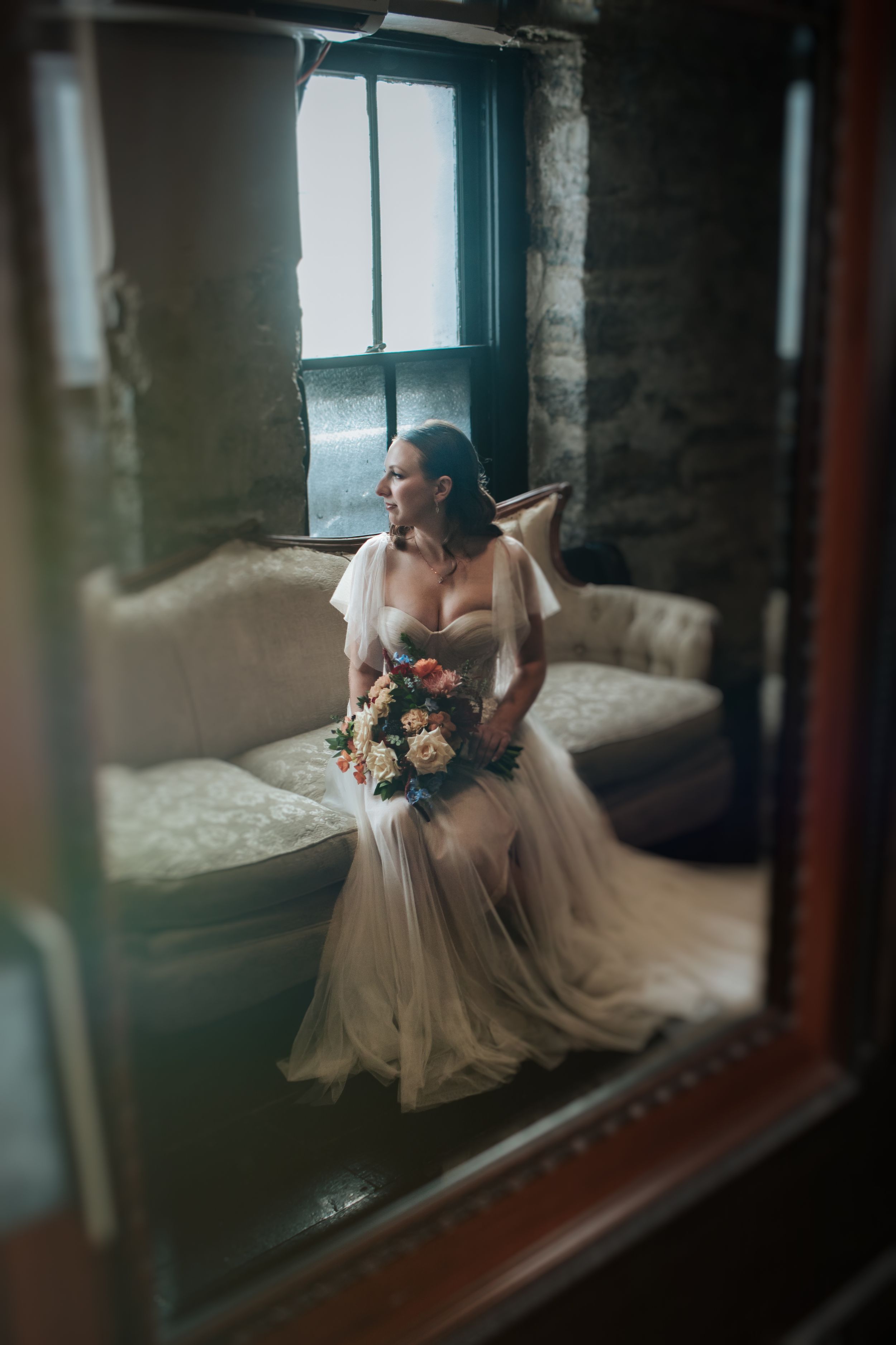 A bride in a flowing tulle gown sits by a window holding a colorful bouquet in a rustic room.