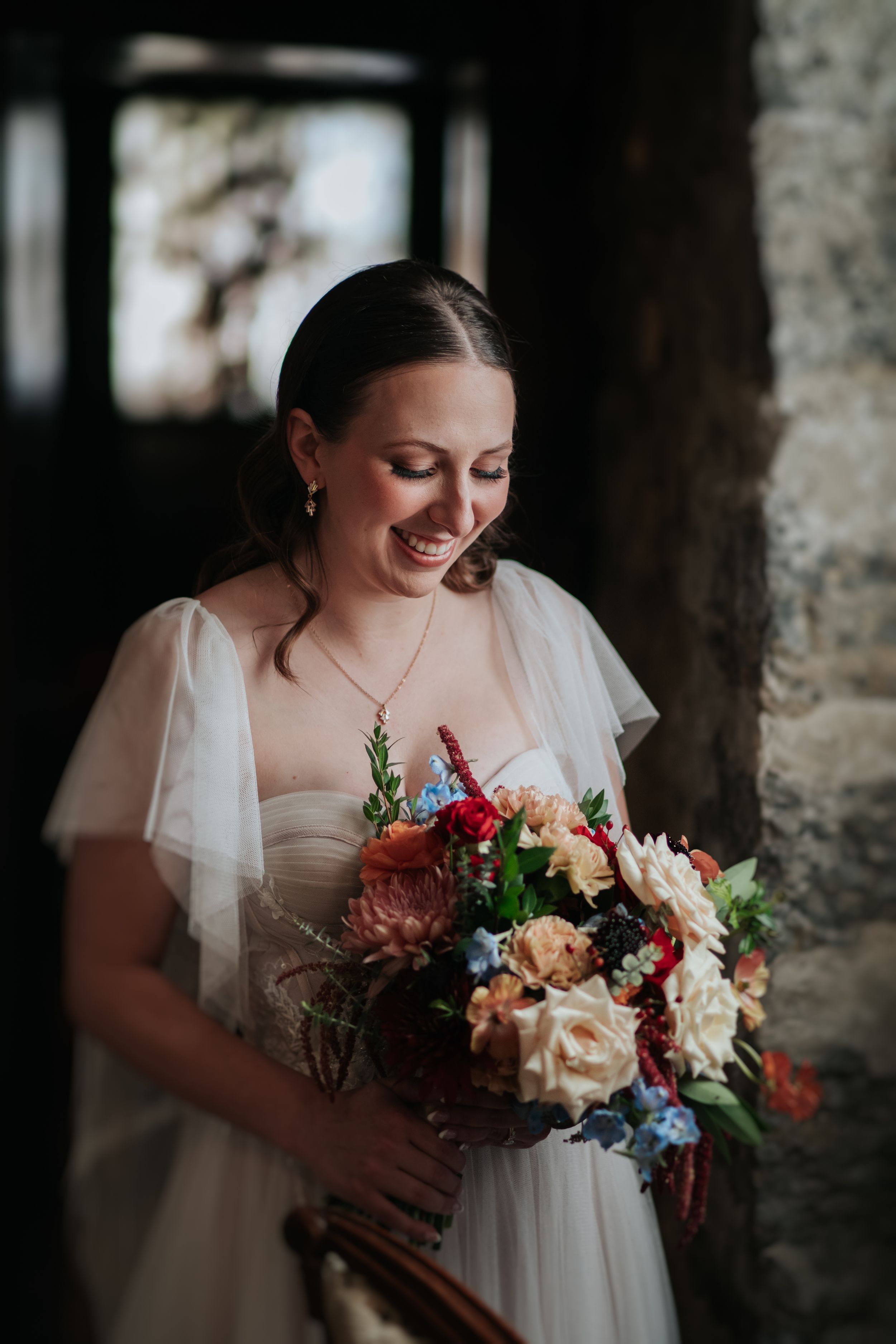 A bride in a flowing white dress holds a vibrant bouquet with burgundy, peach and blue flowers against a rustic stone wall.