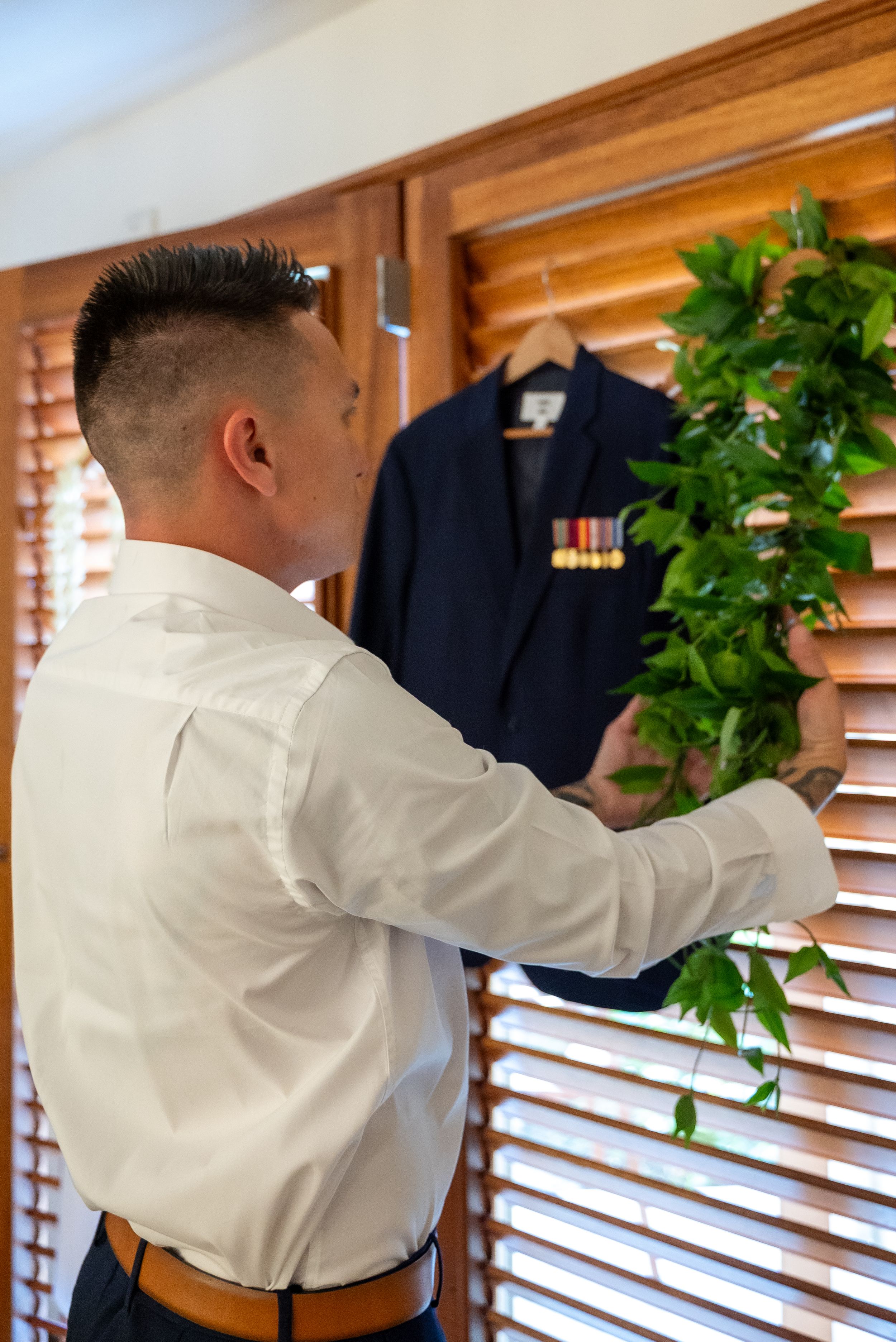 Someone in a white dress shirt and brown pants adjusts window blinds next to a hanging navy uniform with medals.