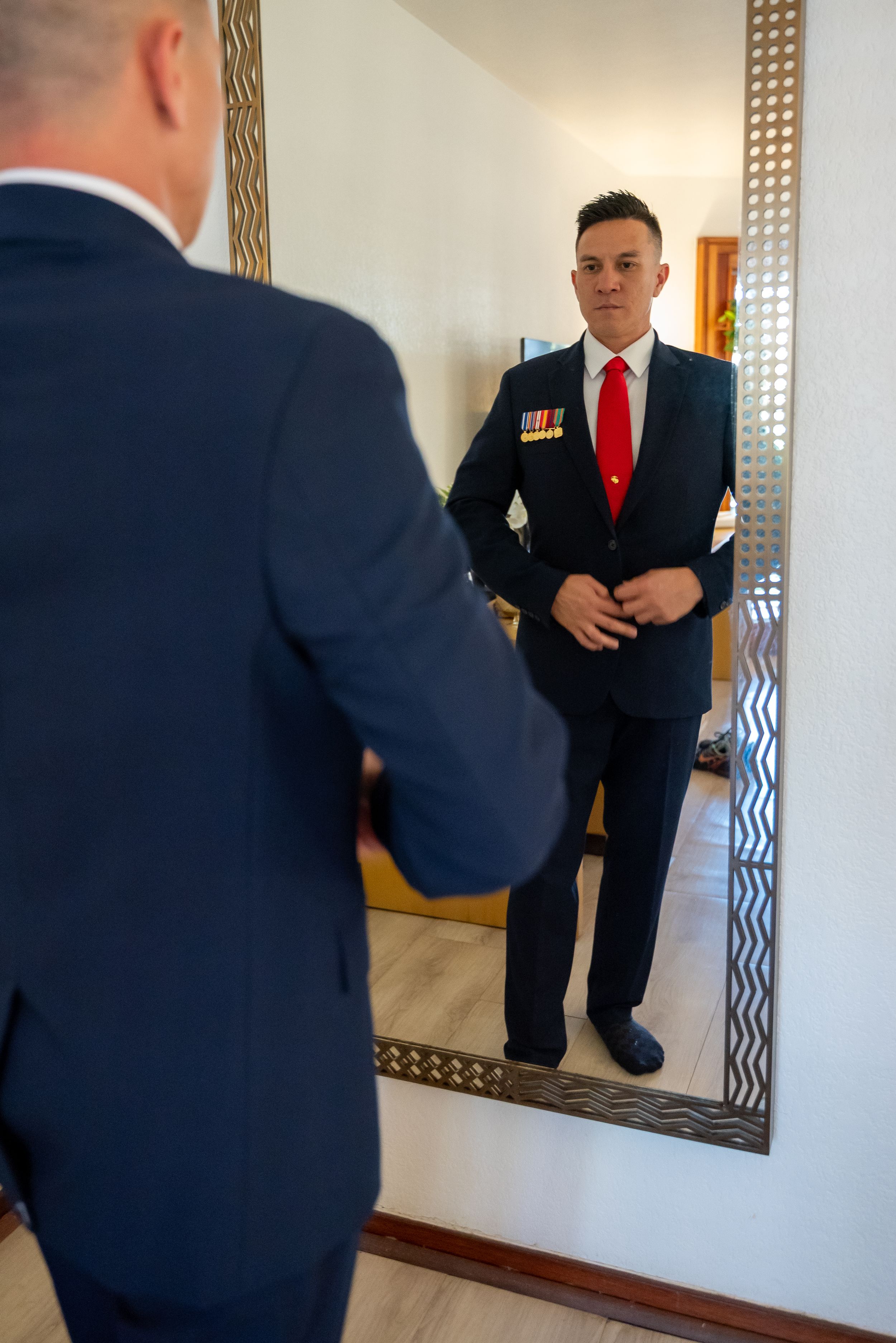 Professional wearing navy suit and red tie adjusts outfit while standing near decorative wall mirror.
