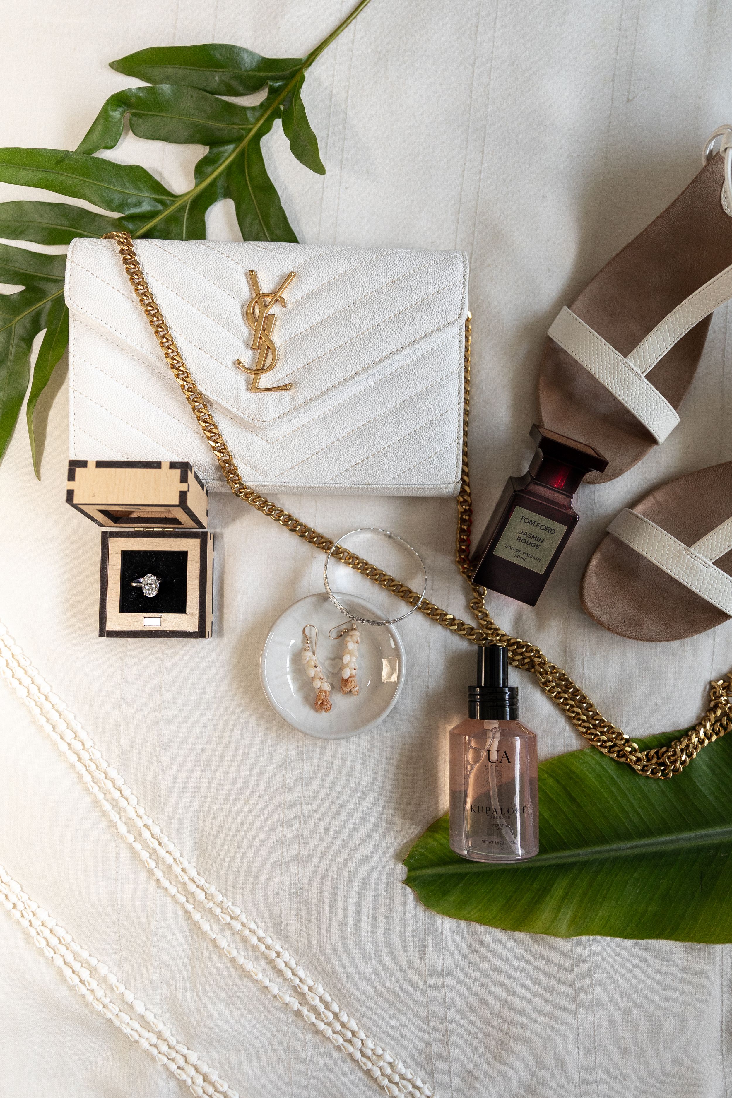 Luxury fashion flatlay with white YSL handbag, jewelry box, perfume bottle and tropical leaves on white background.