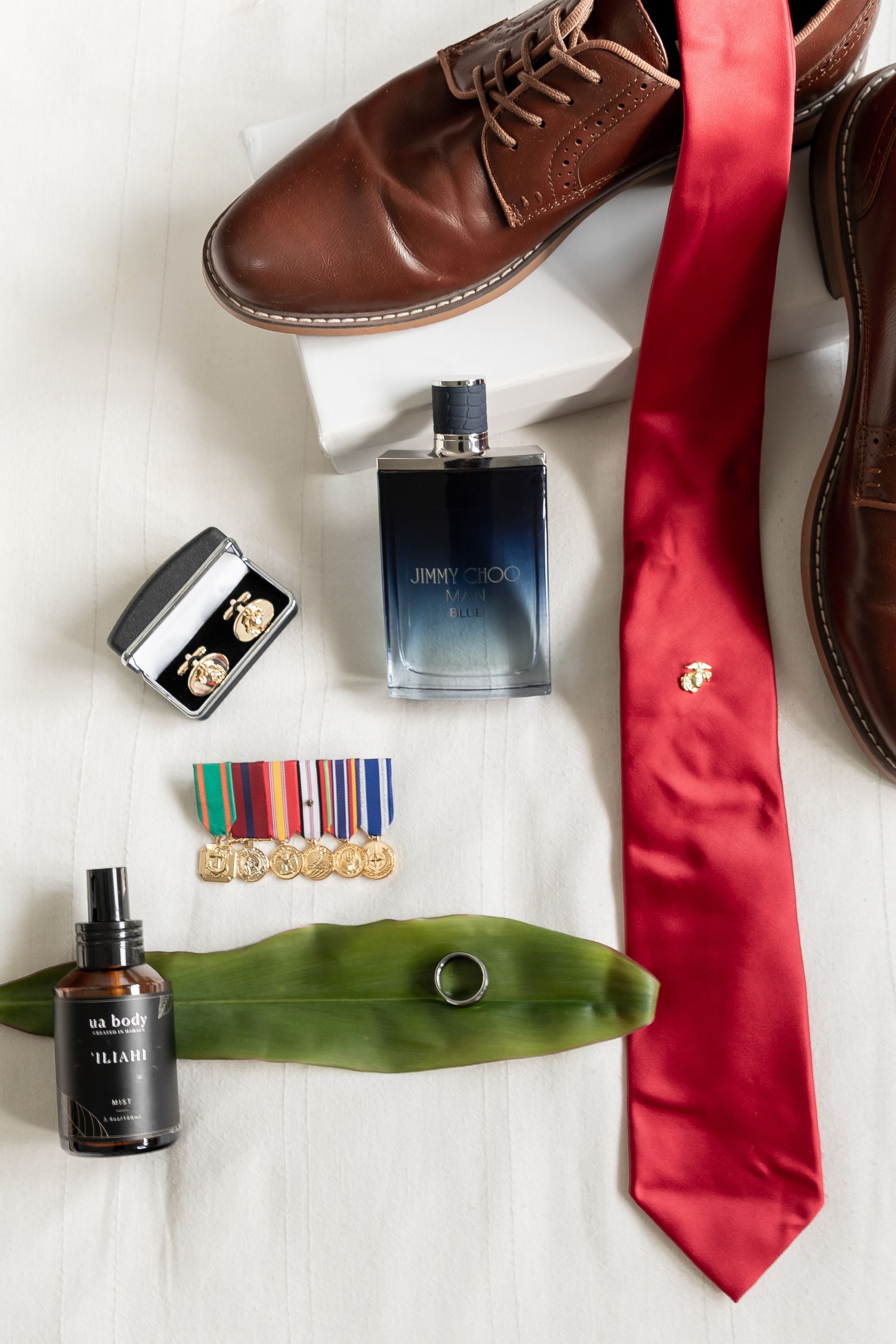 Flatlay of men's accessories including brown leather shoes, red tie, cologne bottles, and grooming items on white surface.