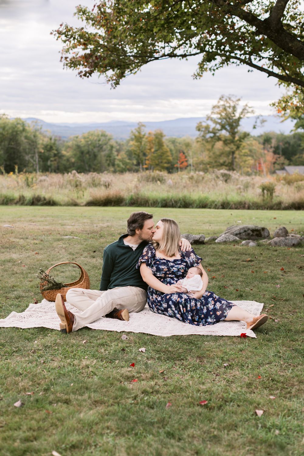 A husband and wife kiss while sitting on a crochet blanket under a tree with mountains in the background while holding their newborn in NH