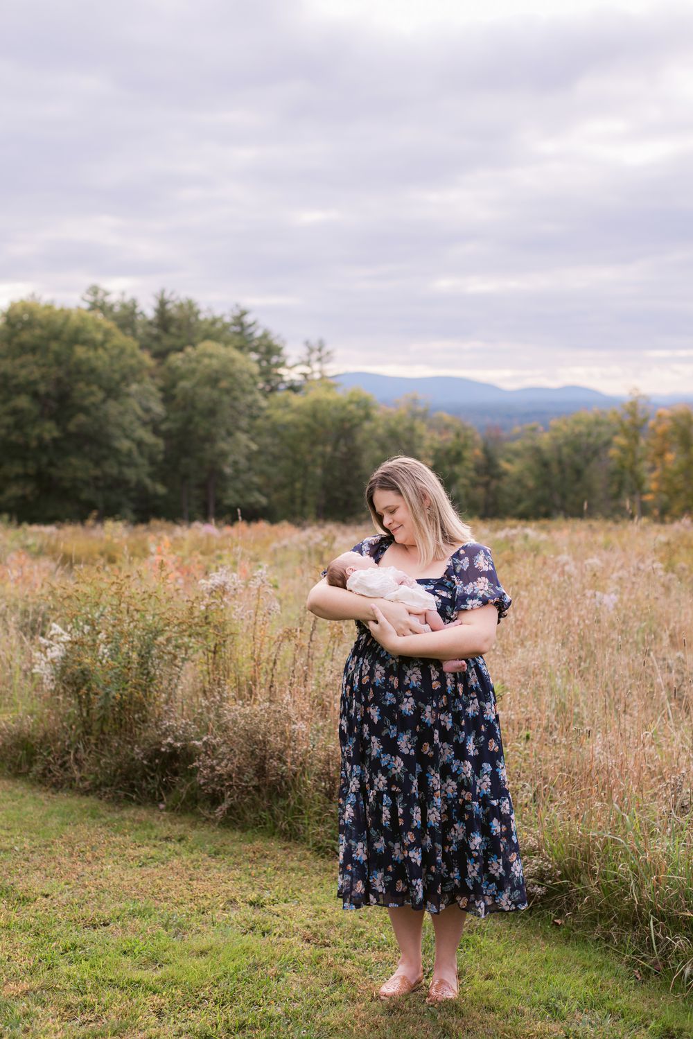 A mother wearing a navy floral dress holds her newborn girl wearing a cream outfit standing next to a golden field in the Lakes Region NH.