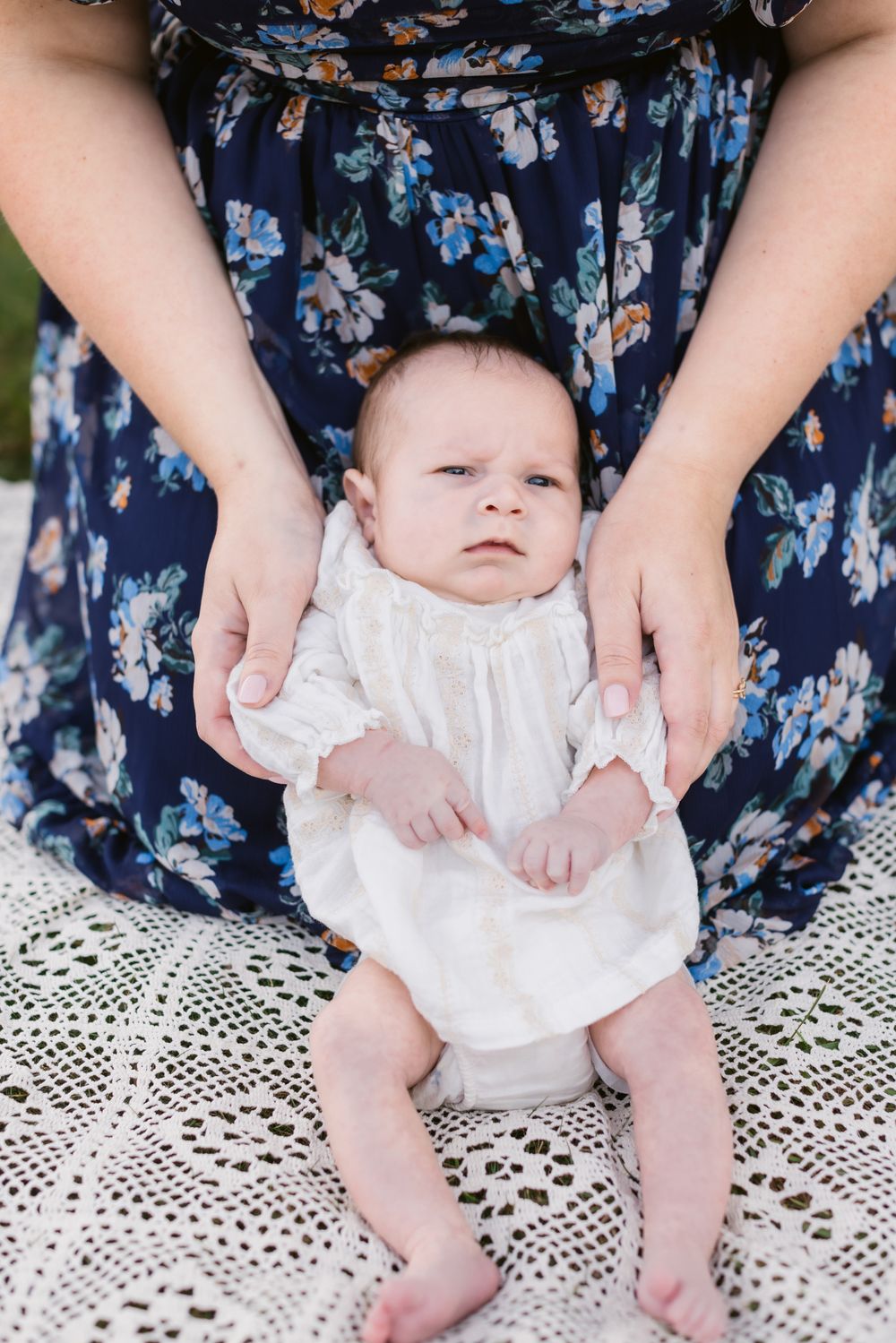 A mother holds her baby girl wearing a cream outfit on a crochet blanket between her knees while kneeling for a newborn shoot in Tamworth NH