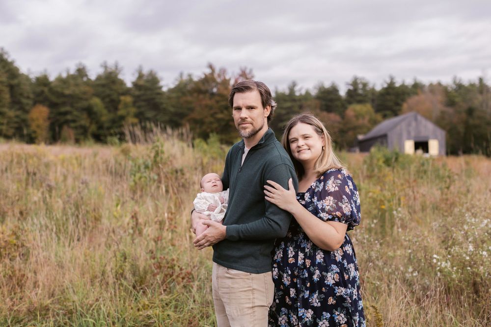 A mother wearing a navy floral dress embraces her husband wearing forest green who holds their newborn during a shoot at Whiteface Hallow NH