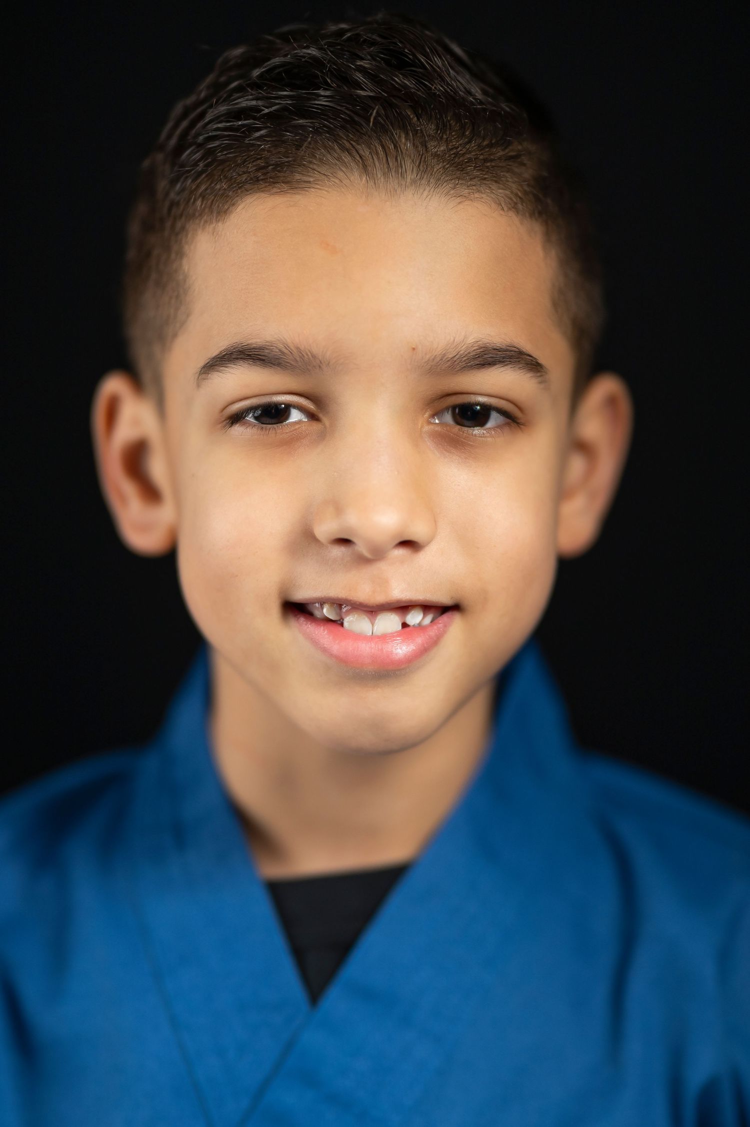 Smiling young boy wearing a blue martial arts uniform against a black background.