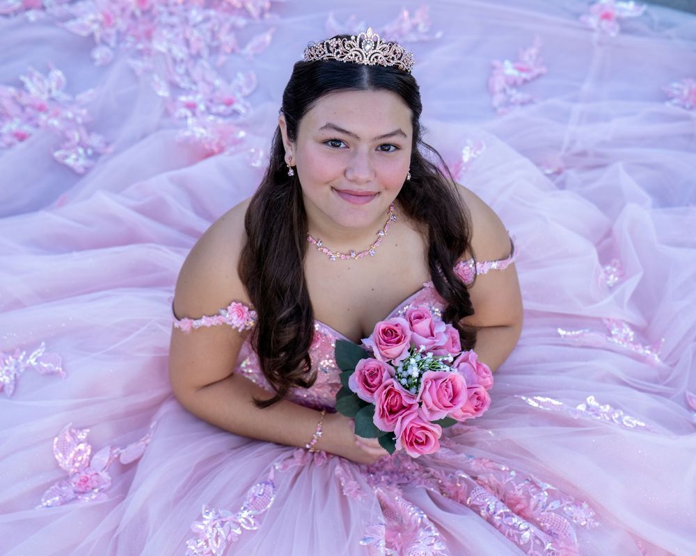 A person in a pink quinceañera or prom dress poses with pink roses against a floral backdrop.