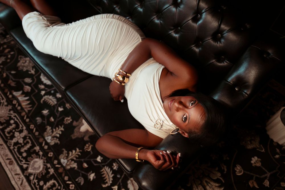 A young Black woman in a white dress and gold jewelry reclines elegantly on a black tufted leather sofa.