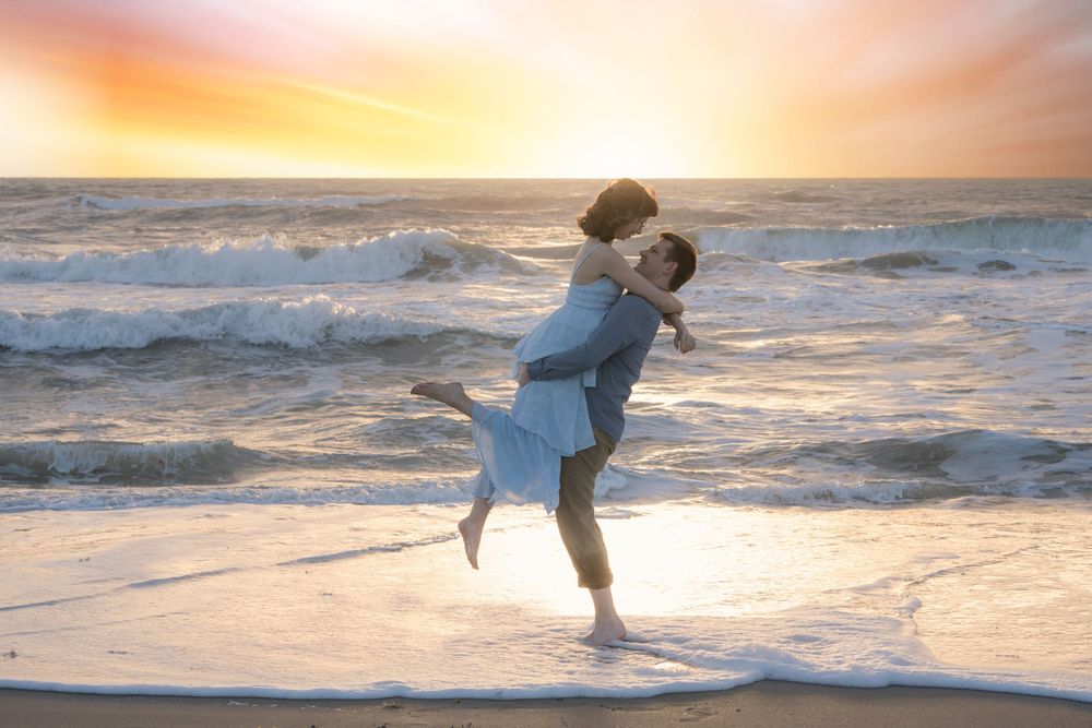 Couple embracing and kissing on a beach at sunset, waves crashing behind them in a romantic seaside moment.