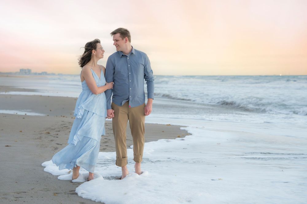 Couple walking hand-in-hand on a beach at sunset, waves washing over their feet, woman in blue dress.