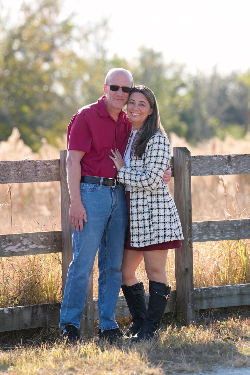 Couple poses together by rustic wooden fence during golden hour sunset in countryside setting.