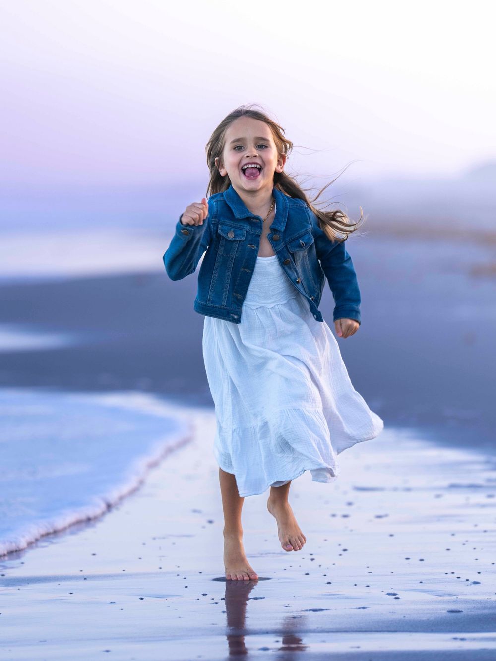 Young girl running and playing joyfully on beach in white dress and denim jacket.