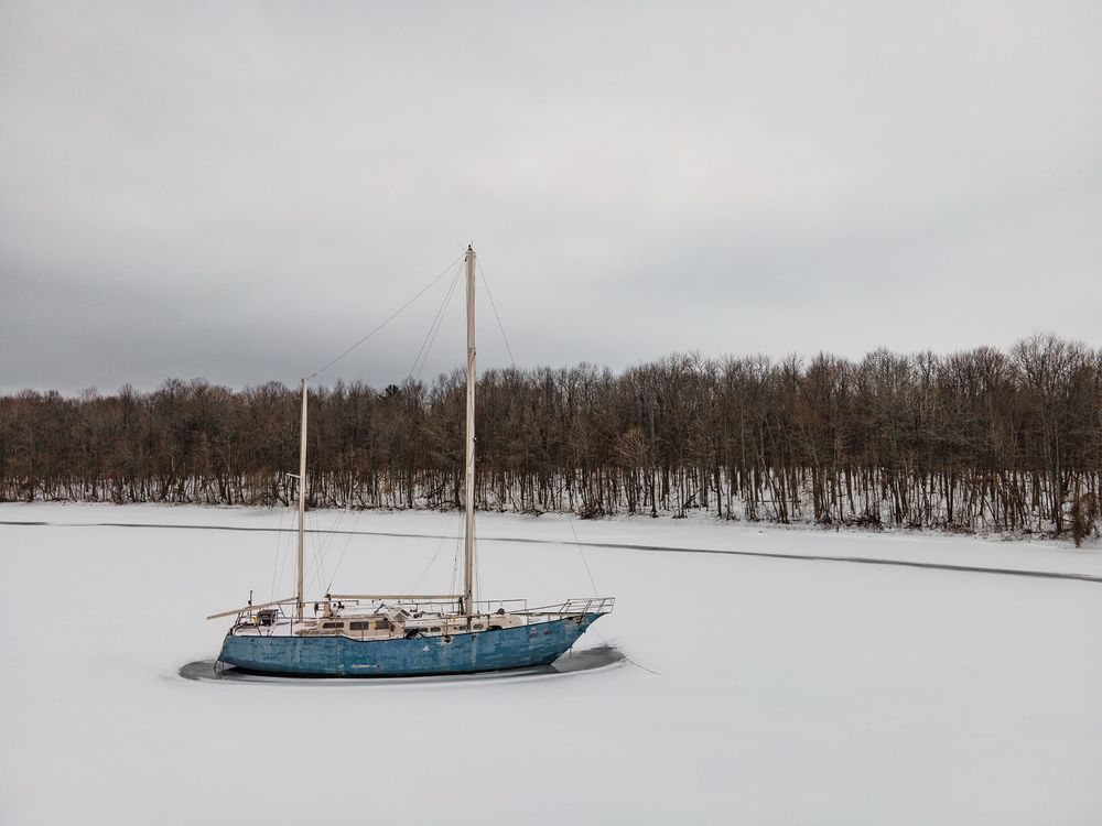 Snow-covered shoreline with bare trees and a blue sailboat moored in the frozen waters.