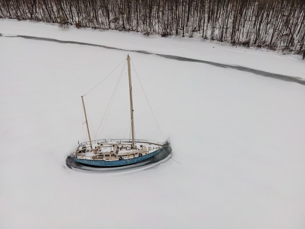 Aerial view of a lone sailboat anchored in a frozen winter harbor surrounded by snow-covered landscape.