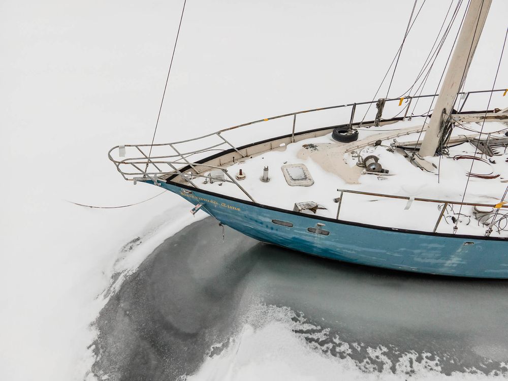 Snow-covered sailboat moored in winter waters with white deck and blue hull.