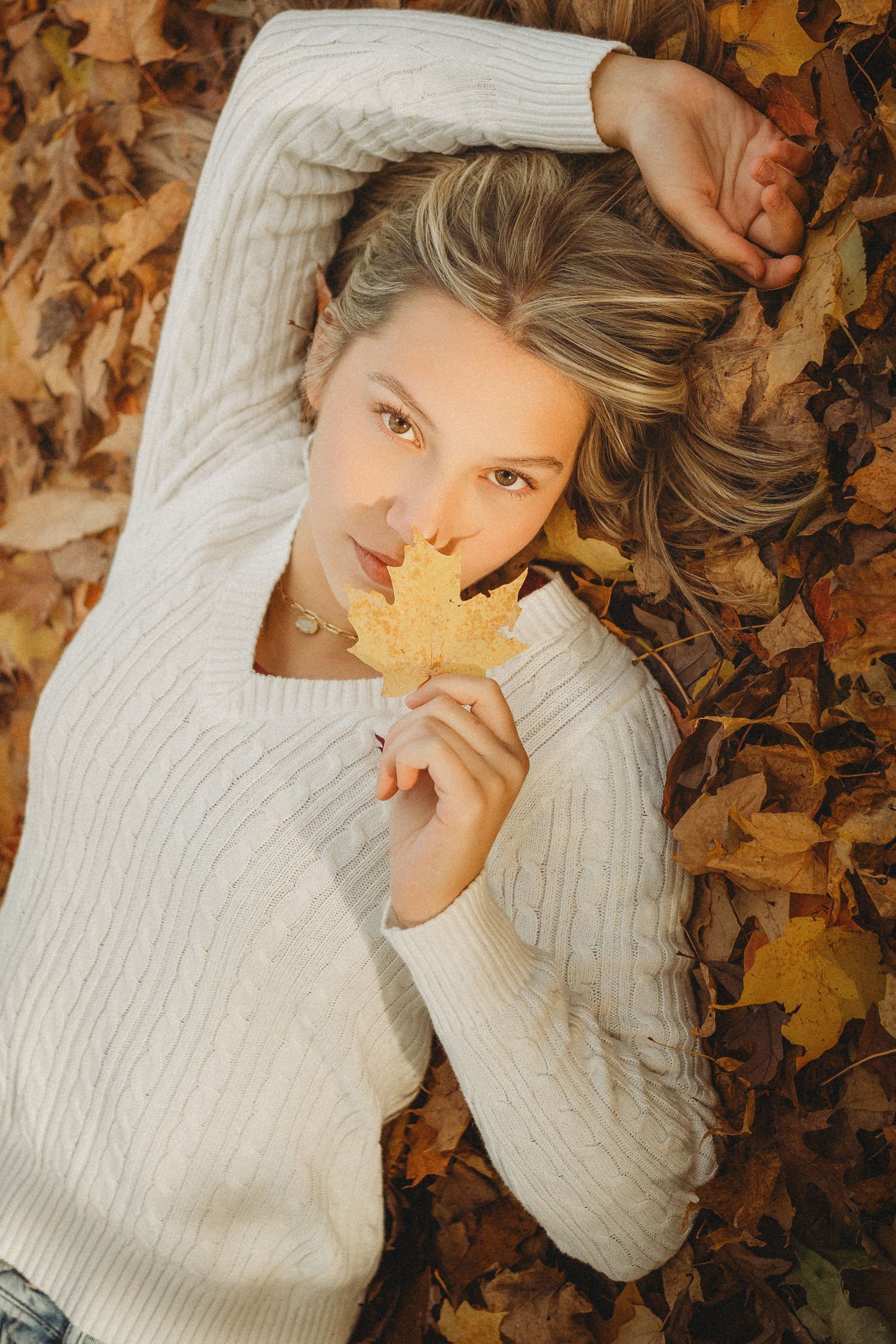Blonde teen girl lying in autumn leaves holding maple leaf near face wearing white sweater.