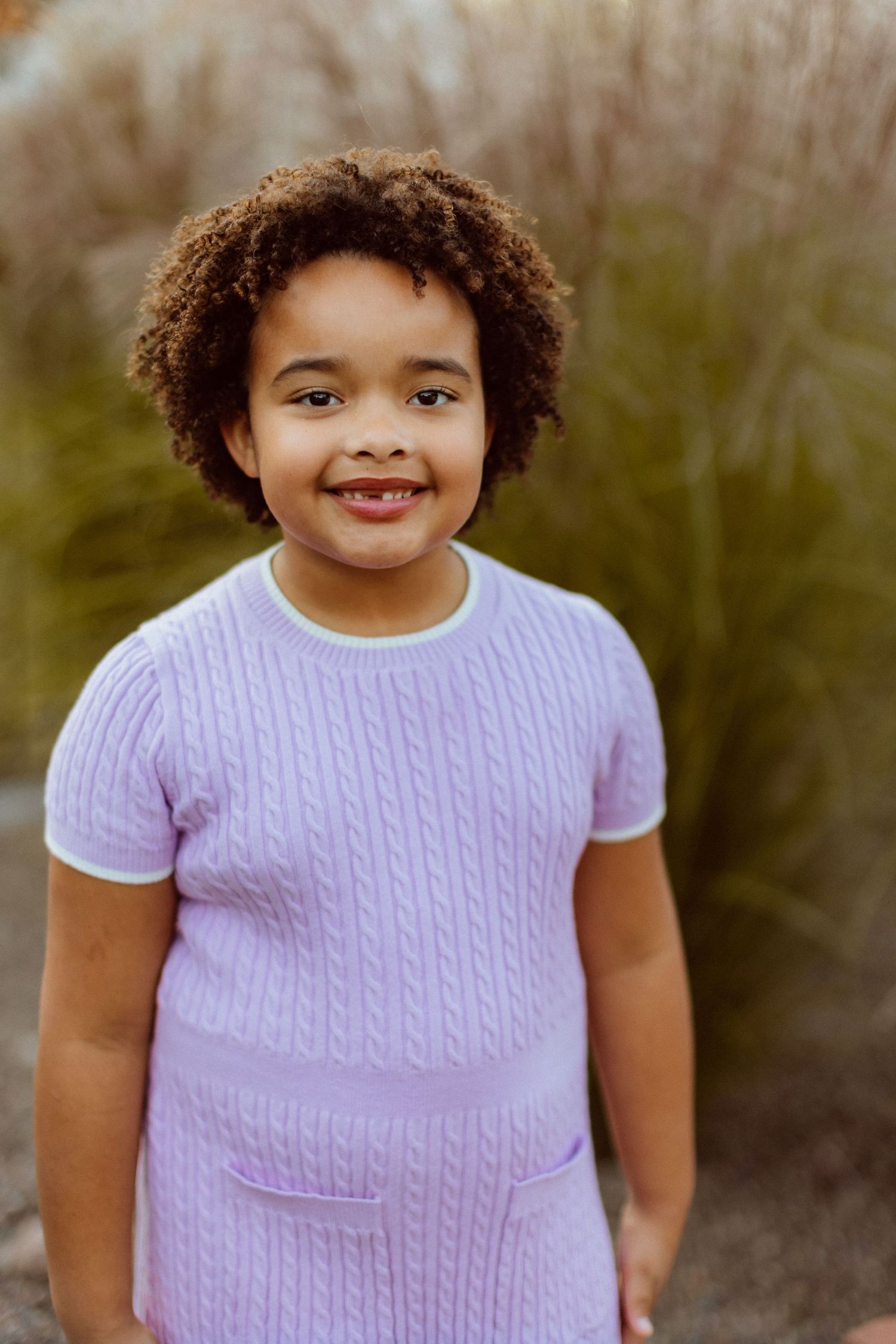 A smiling girl poses for the camera in a lilac dress in front of tall prairie grasses at her backyard family photo session.