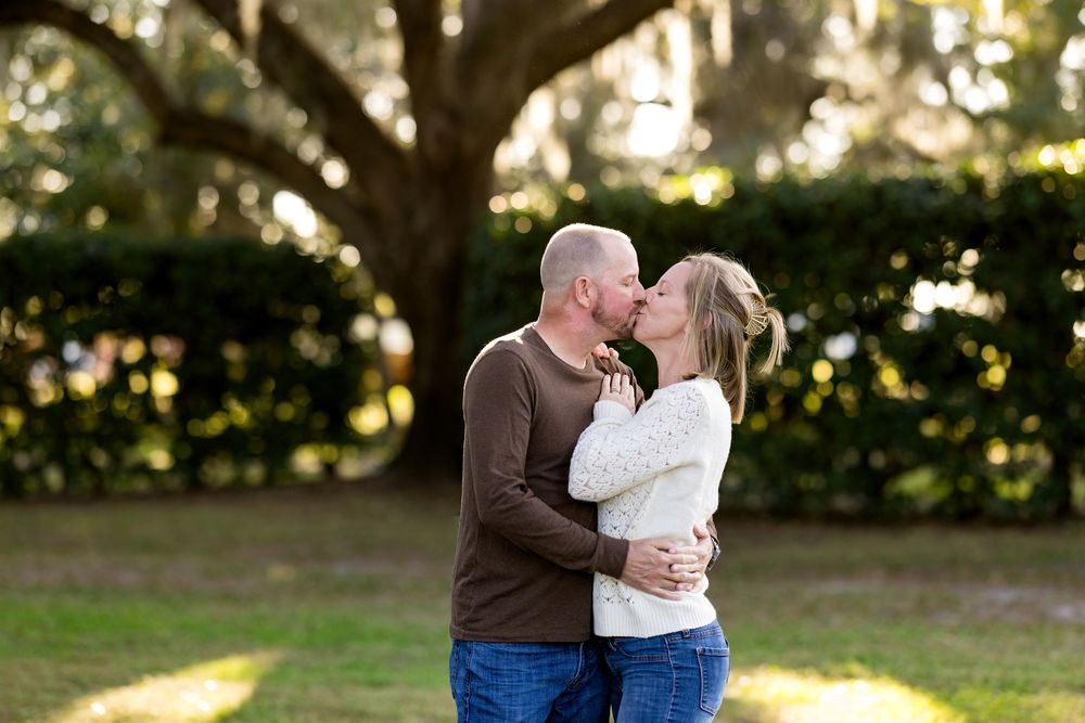 A couple embraces in a romantic outdoor photoshoot with soft sunlight filtering through trees in the background.