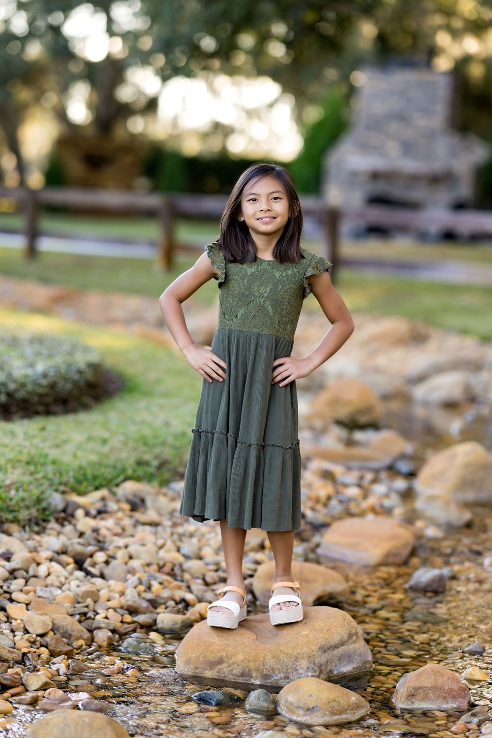 A child in a green dress stands on rocks near a stream in a park setting during golden hour.