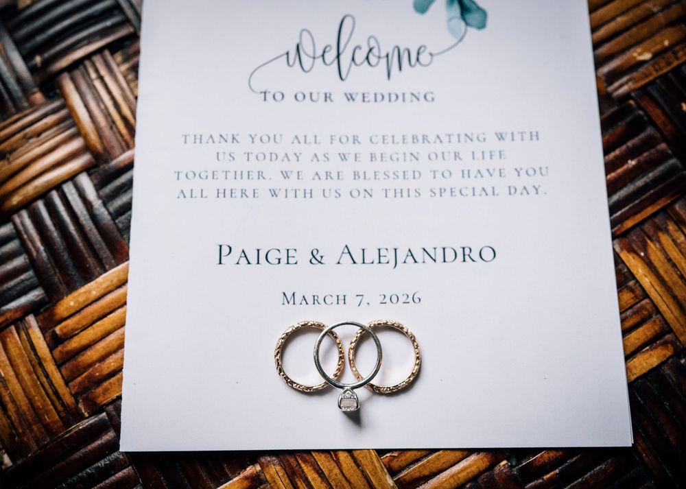 Wedding rings and invitation details photographed on a woven bamboo surface with elegant calligraphy text.