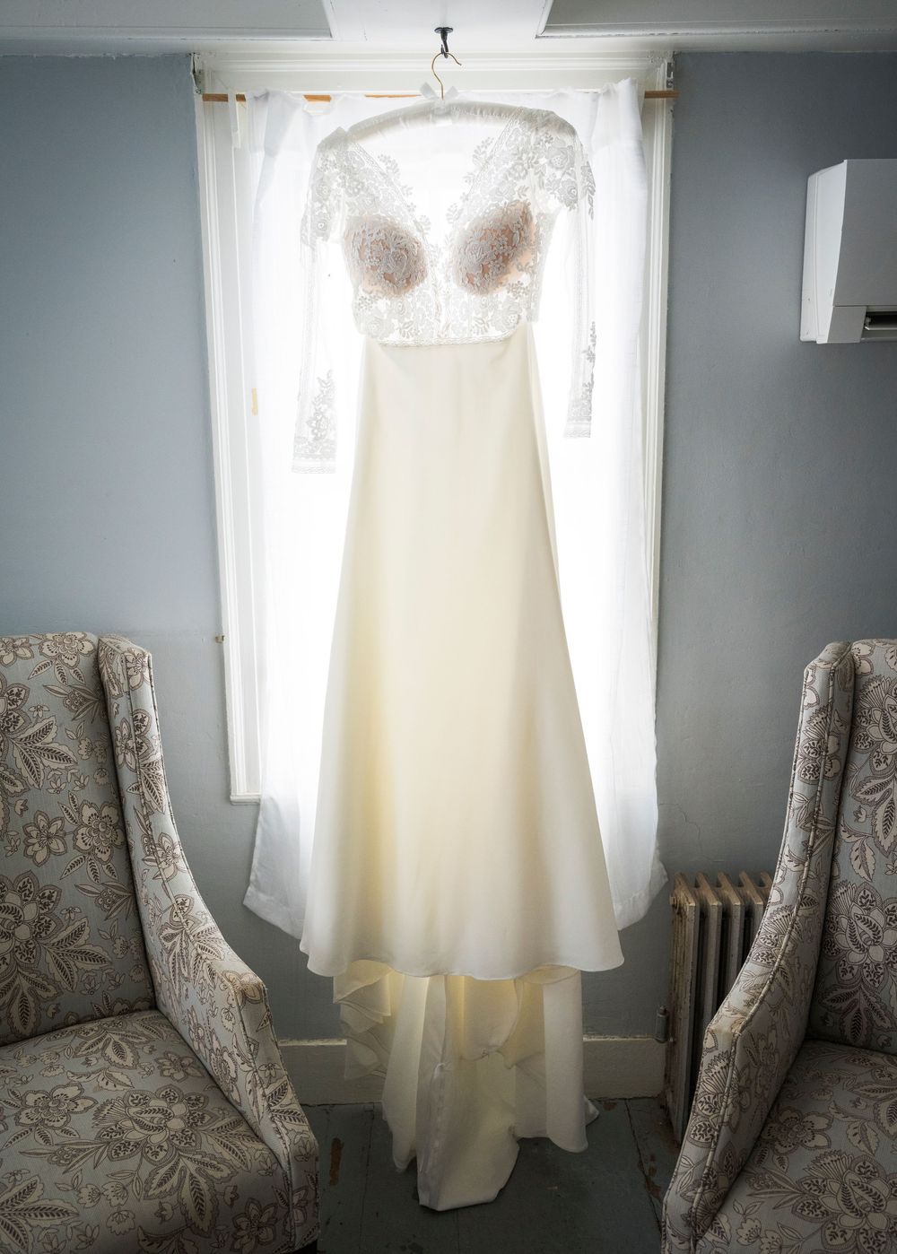 White wedding dress hanging in a room with gray walls and patterned accent chairs, illuminated by natural light.
