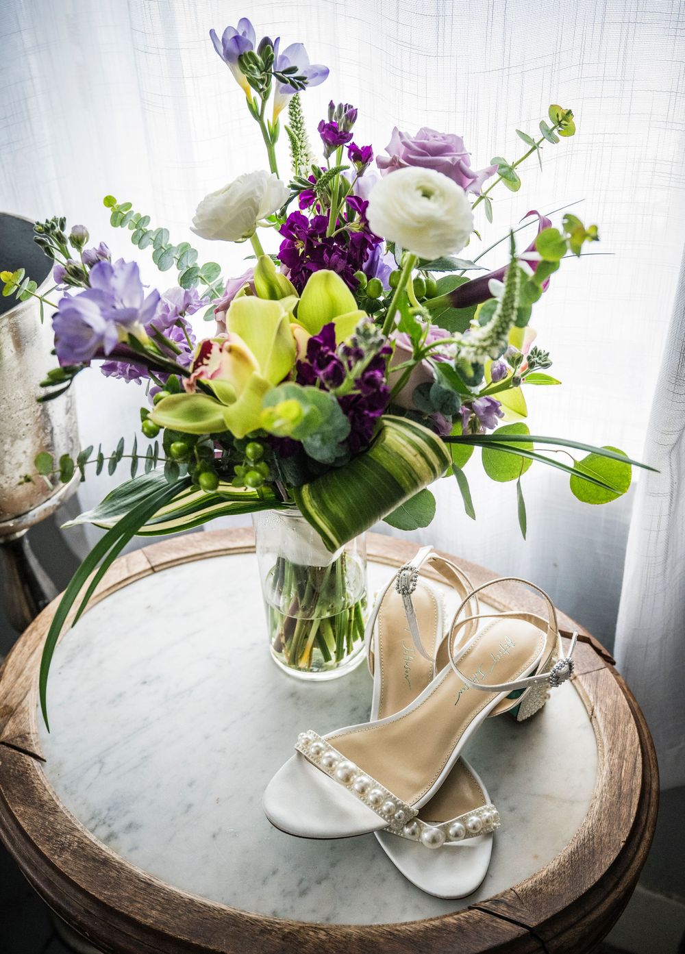 Elegant floral arrangement with purple and white flowers sits atop a rustic wooden surface near a window.