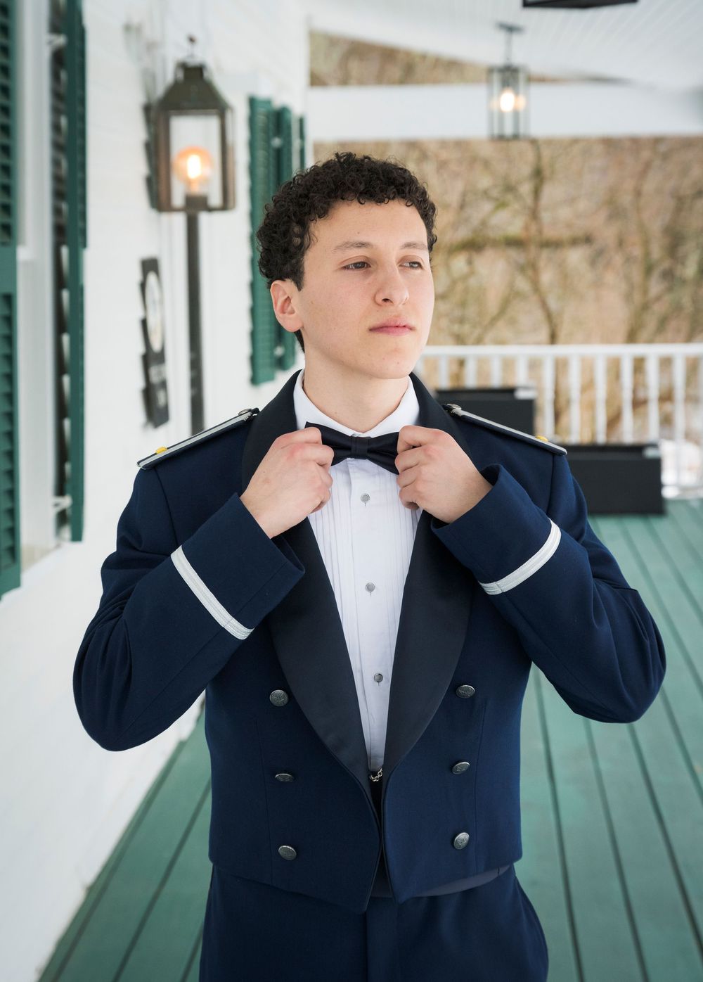 A person adjusts their bow tie while wearing a formal navy tuxedo on a porch with white columns and green flooring.