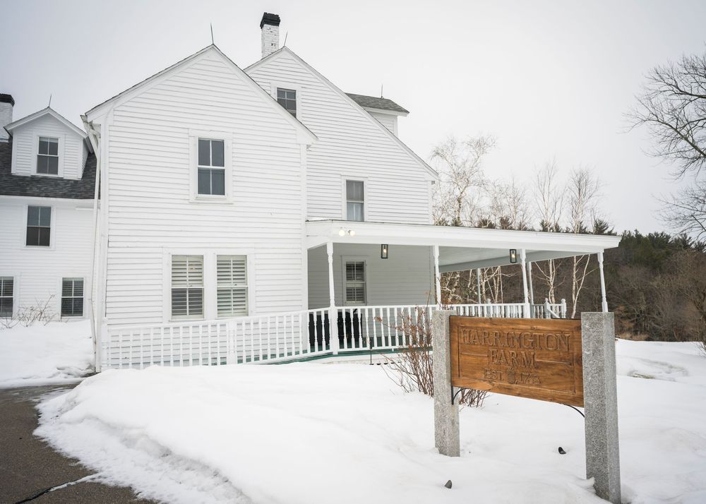 Snow covers the ground outside a white farmhouse with a wooden fence and bare trees in winter.