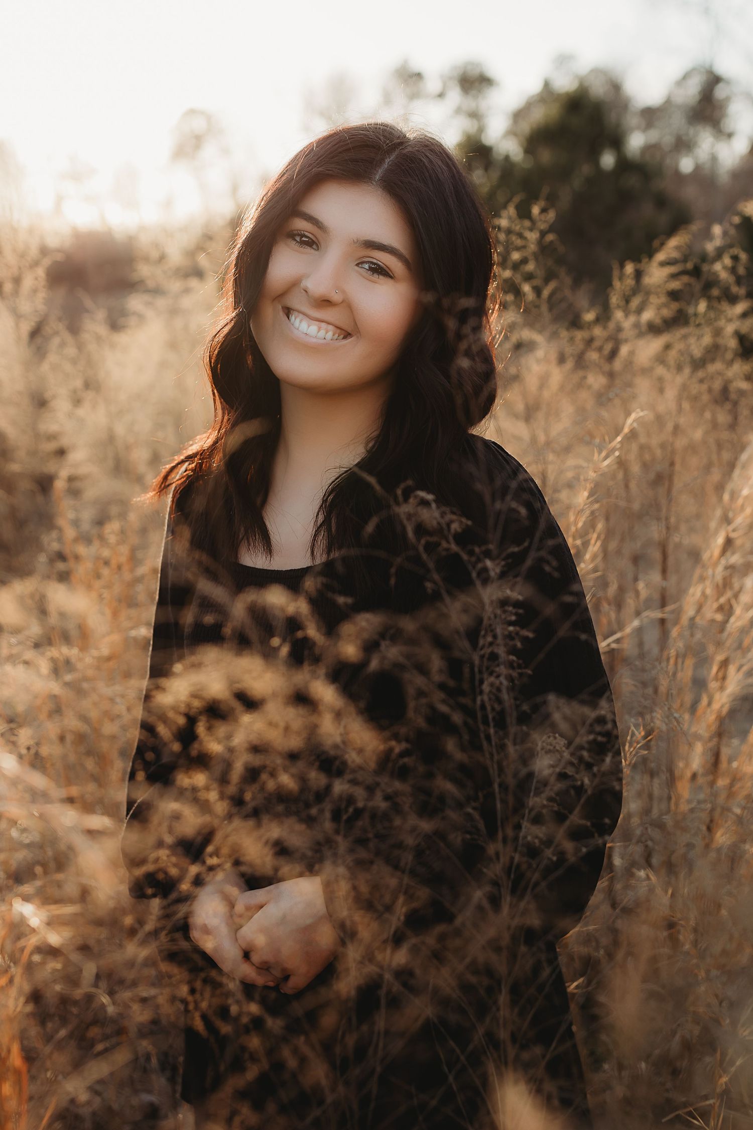 Portrait photo of someone smiling in a black sweater against golden sunlit dried grass during golden hour.