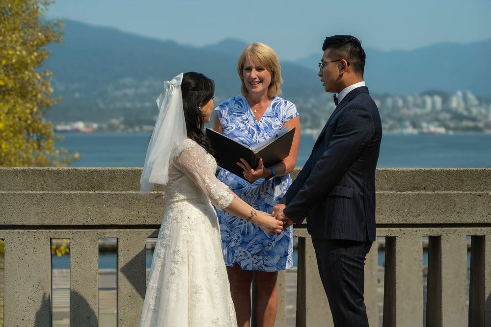 An outdoor wedding ceremony takes place on a stone terrace overlooking mountains and ocean on a sunny day.