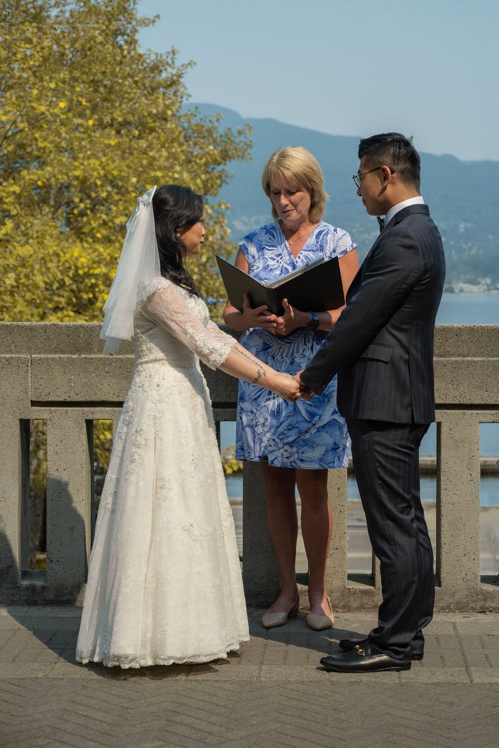 A wedding ceremony takes place on a scenic overlook with mountains and water in the background.