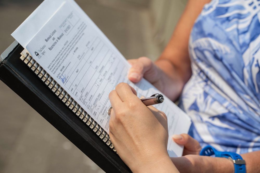 Someone writes in a spiral notebook with a blue pen while wearing a blue and white patterned shirt.