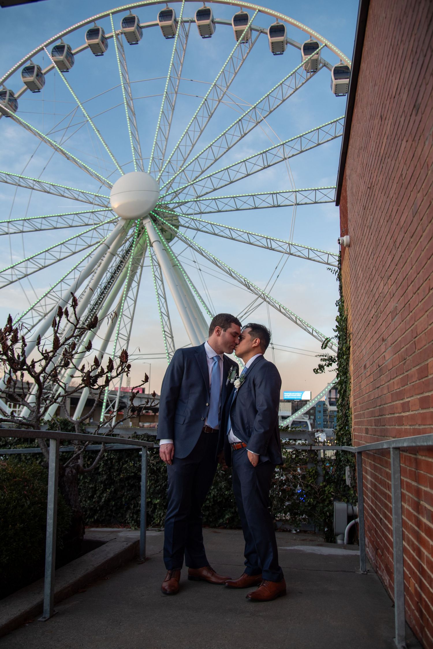 A romantic couple shares an intimate moment in front of a large Ferris wheel at sunset.