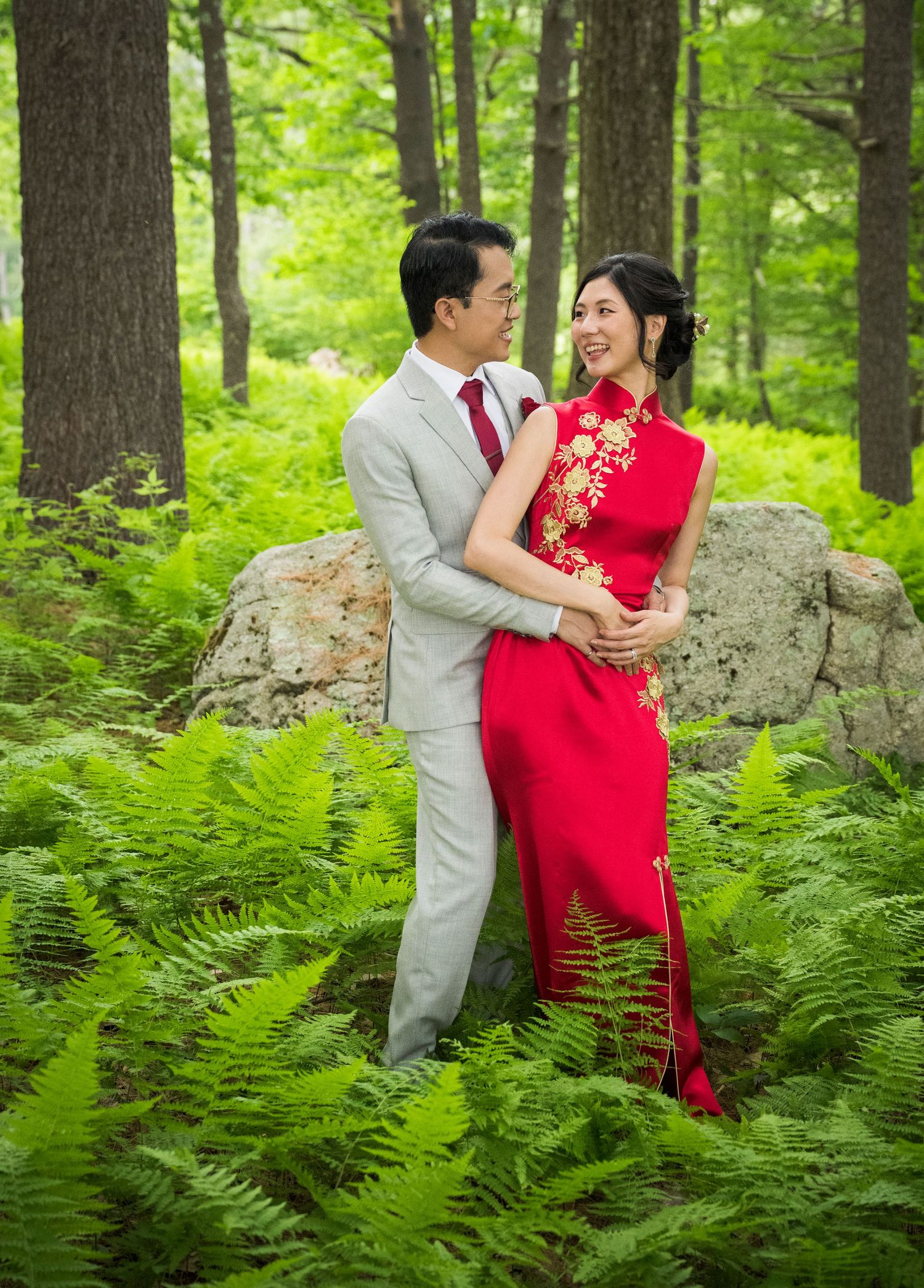 A couple in traditional wedding attire poses romantically among lush green ferns in a forest setting.