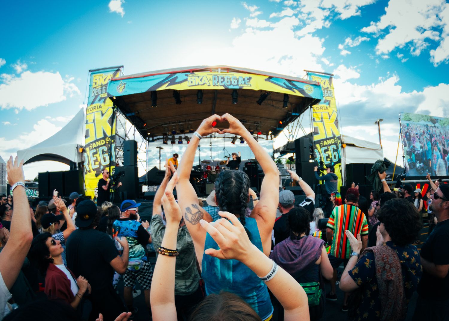 Silhouetted crowd raises hands in unison against bright blue sky during an outdoor music festival or concert.